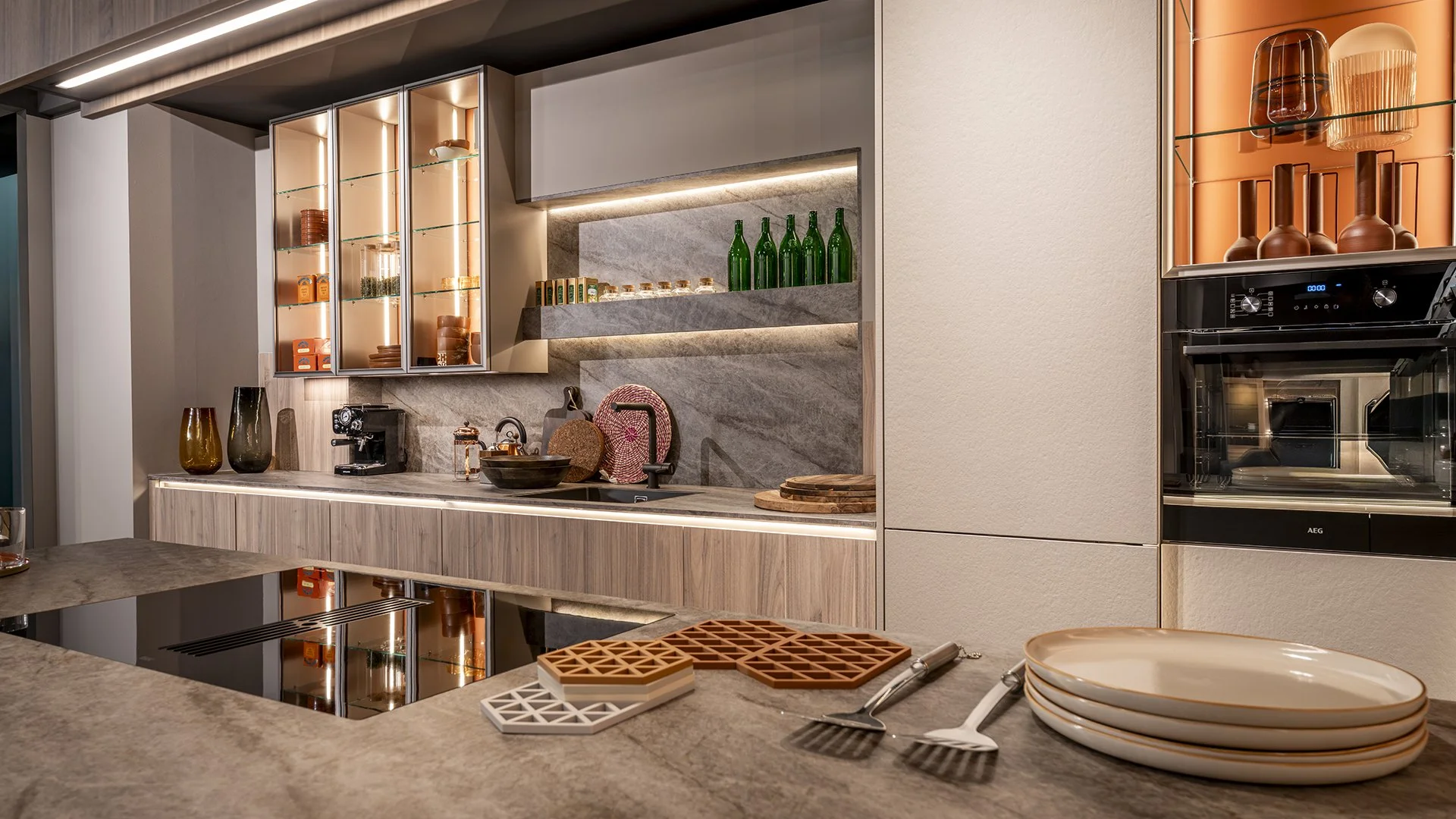 Modern kitchen with grey and beige cabinets, open shelves with green bottles and glassware, beige countertops, and a built-in oven. Decorative vases, a coffee machine, and various kitchen utensils are on the counter. There are stacks of plates and baking tools on the foreground counter.