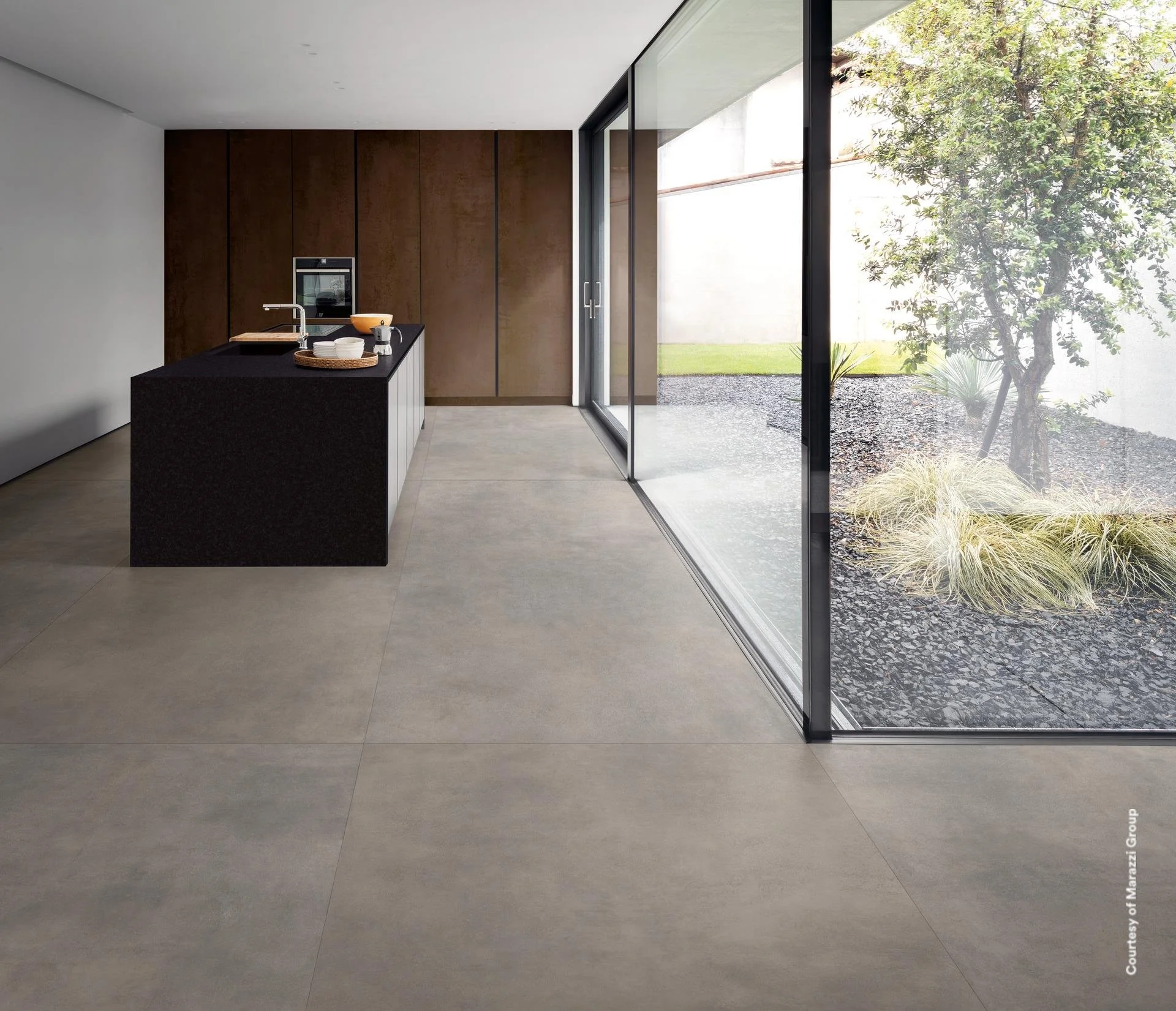 Modern kitchen with dark countertop island, white plates, a brown bowl, and a silver kettle, next to a large glass sliding door showing a small outdoor garden with grass, gravel, and a tree. Marazzi premium surfaces. German Kitchens bespoke.