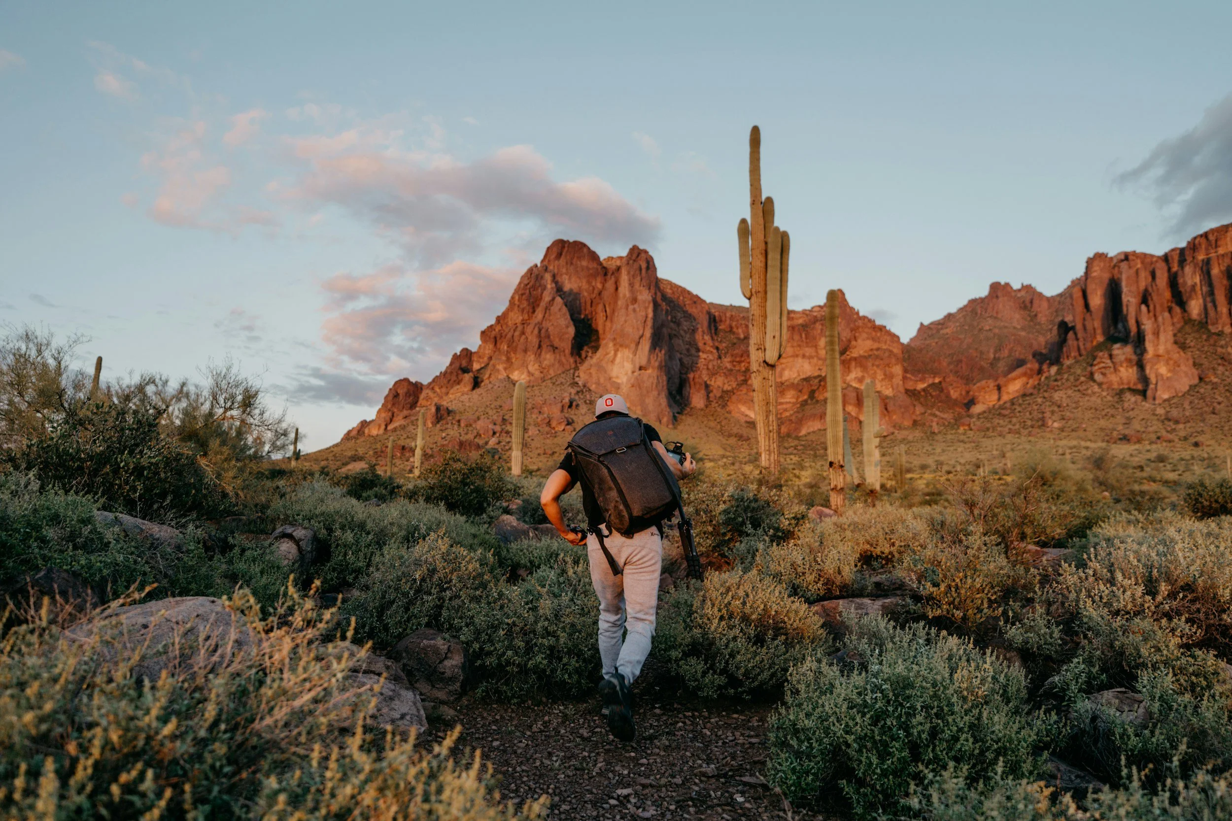 An older person with a backpack hiking through the Sonoran Desert landscape with saguaro cacti and red-rock mountains at sunset.