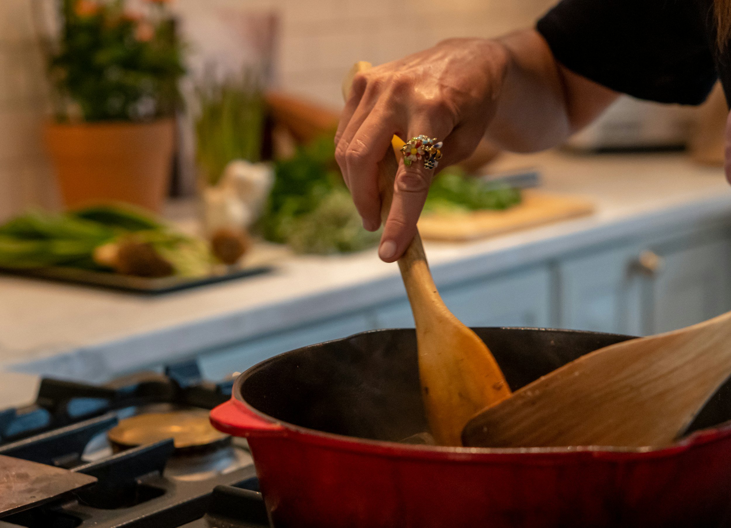 A person's hand stirring food in a red bowl on a stovetop with a pot and potted plants in the background.