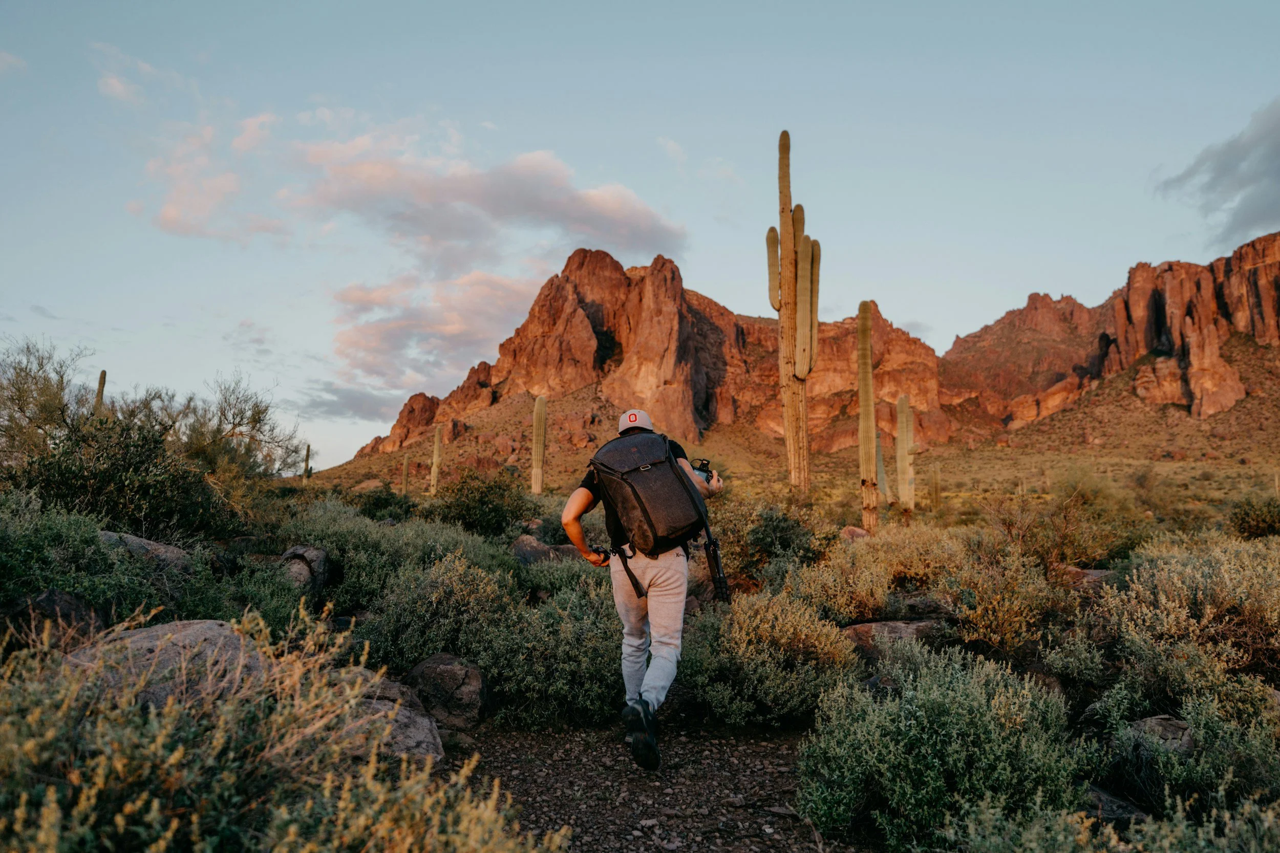 A person wearing a white cap, black backpack, and light-colored pants hiking through desert terrain with Saguaro cacti and red rocky mountains in the background during sunset.