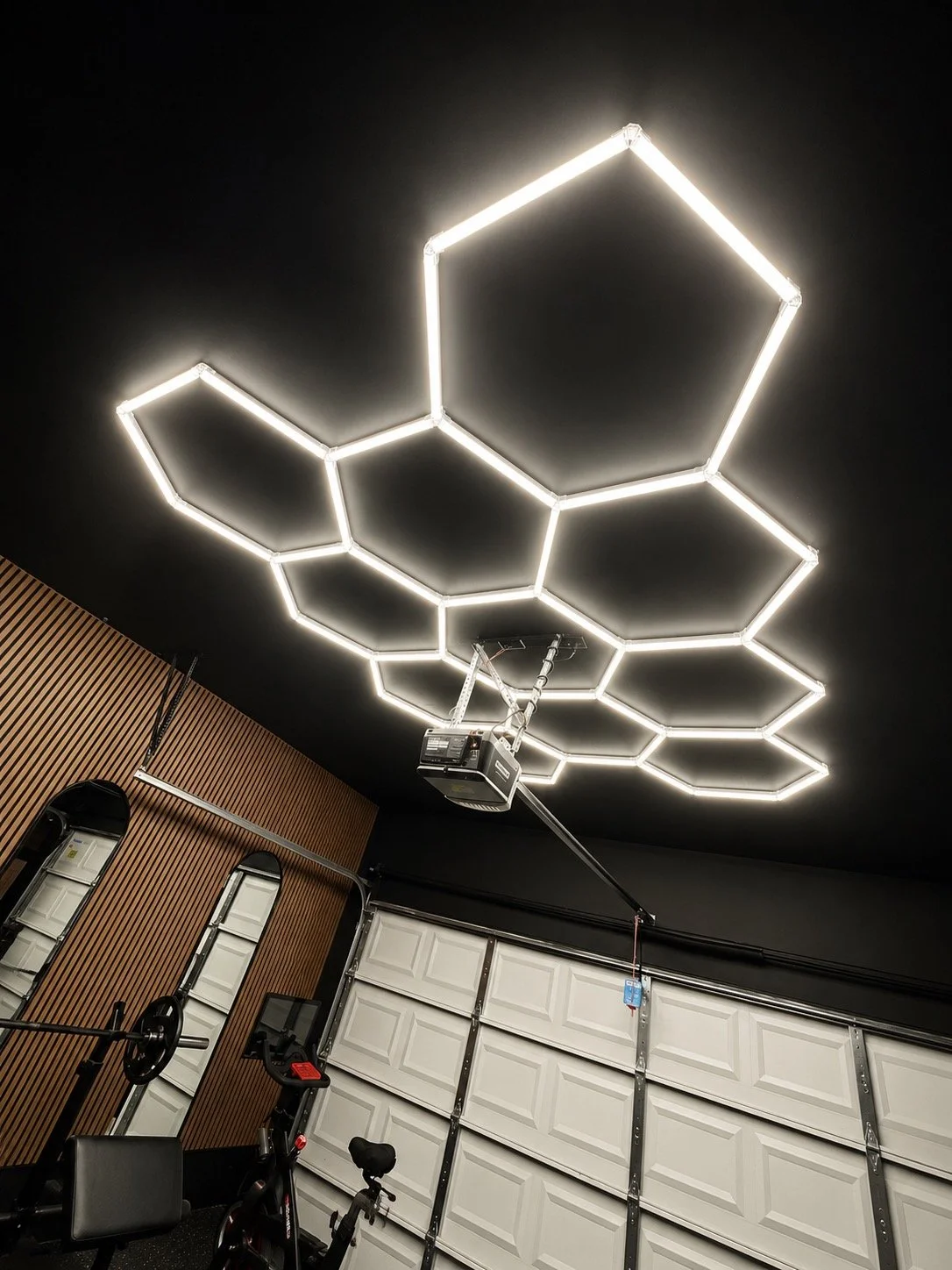 Modern gym with black ceiling featuring geometric LED light fixture and various exercise equipment including a stationary bike and weightlifting barbell.