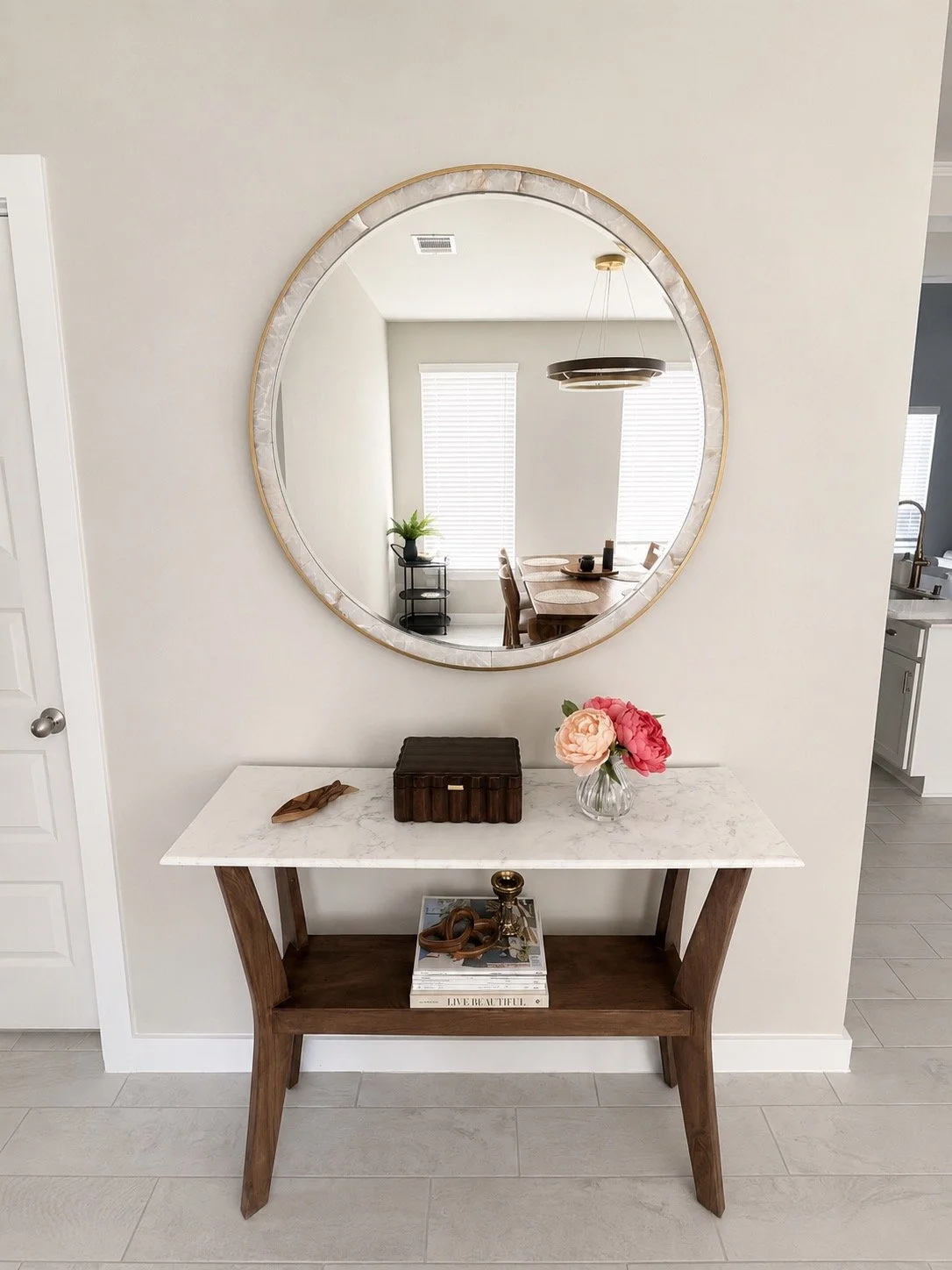 A circular mirror hangs on a white wall above a modern console table with a marble top and wooden legs. The table is decorated with a small glass vase with pink and red flowers, a dark wooden box, and a decorative metallic object. Reflected in the mi