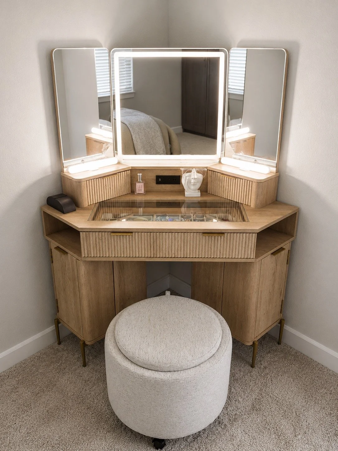 Wooden vanity table with three mirrors, a stool, and decorative items such as a perfume bottle and a white facial mask sculpture. The vanity also has built-in lighting and a glass top compartment, with a bedroom visible in the reflection.