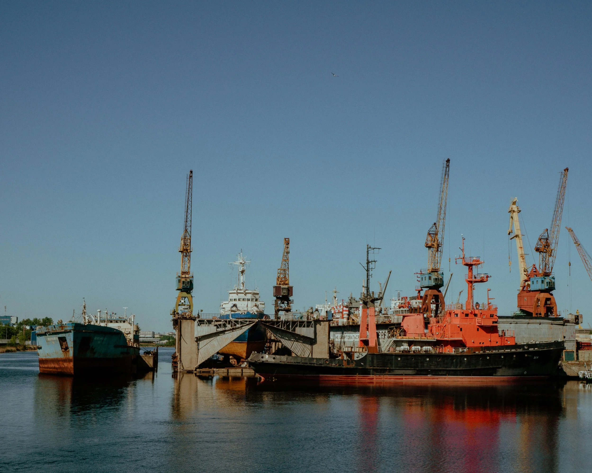Several cranes and ships docked at a port with calm water and a clear sky.