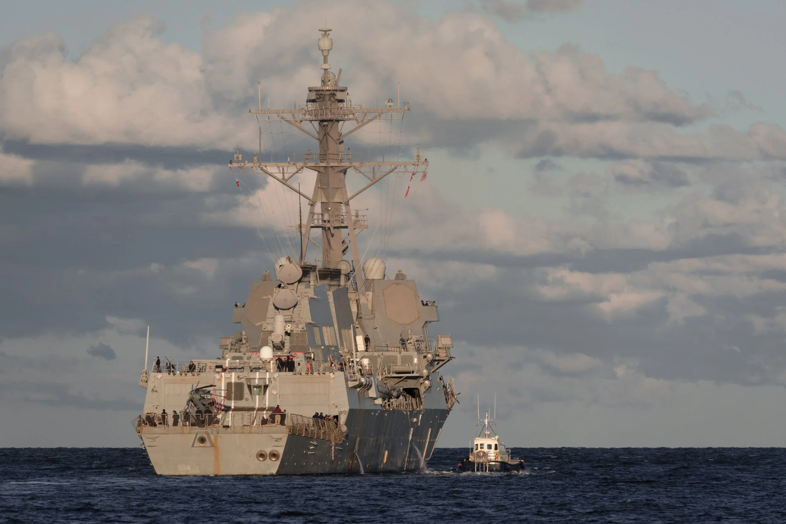 A large naval ship on the water being guided by a small tug boat, with a sky filled with clouds in the background.