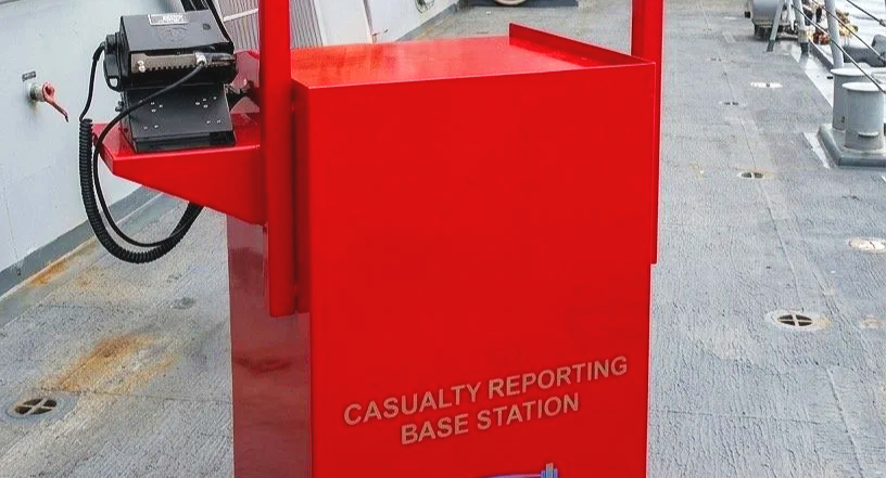 Central Radio Co, Inc.'s red casualty reporting base station with a phone mounted on top, located on a ship or industrial setting with grey flooring and metal structures.