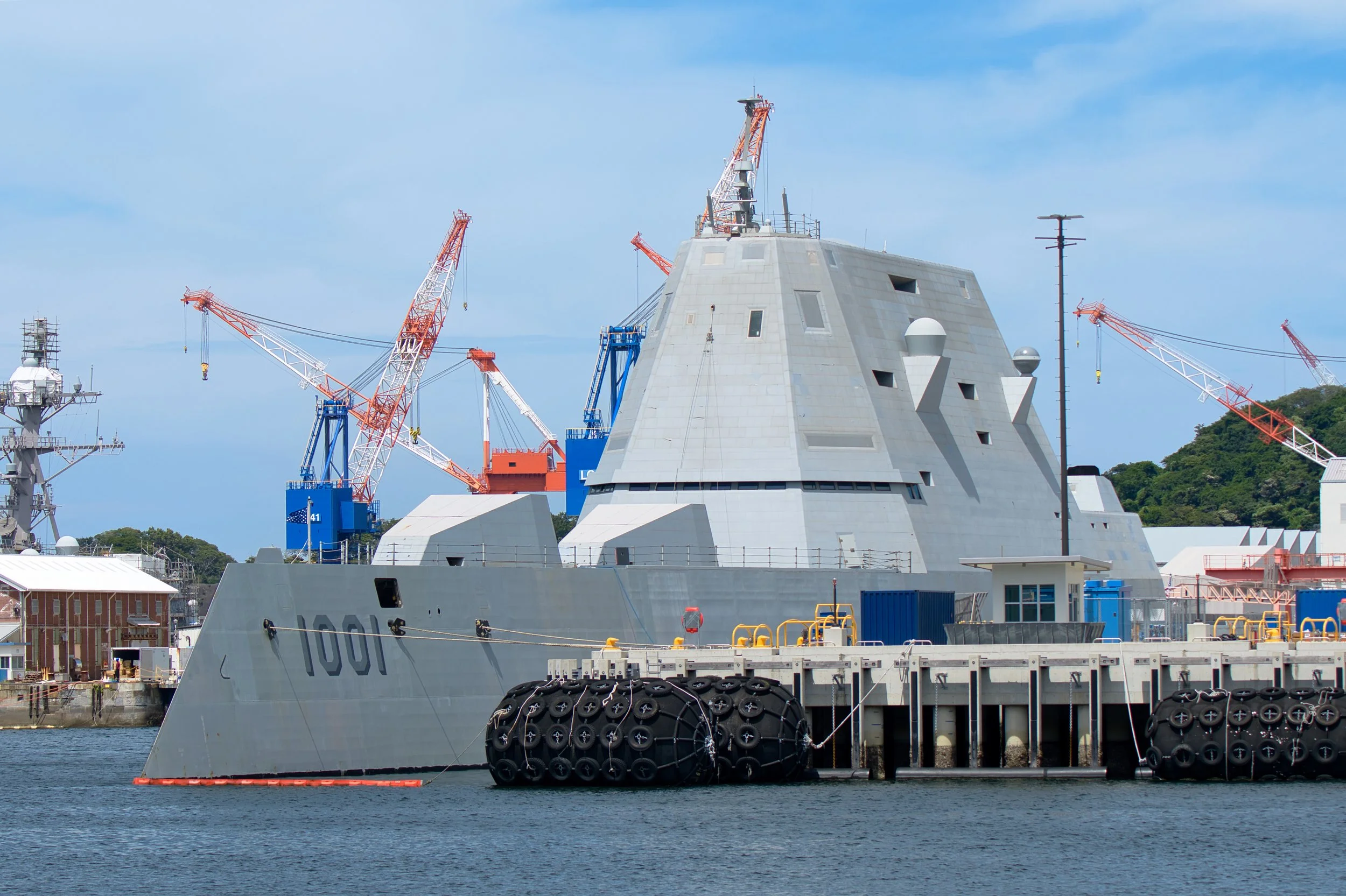 A large naval ship docked at a harbor with multiple cranes around it, the ship is gray with the number 1001 visible on its side, and has various equipment on its deck, with a background of blue sky and some greenery.
