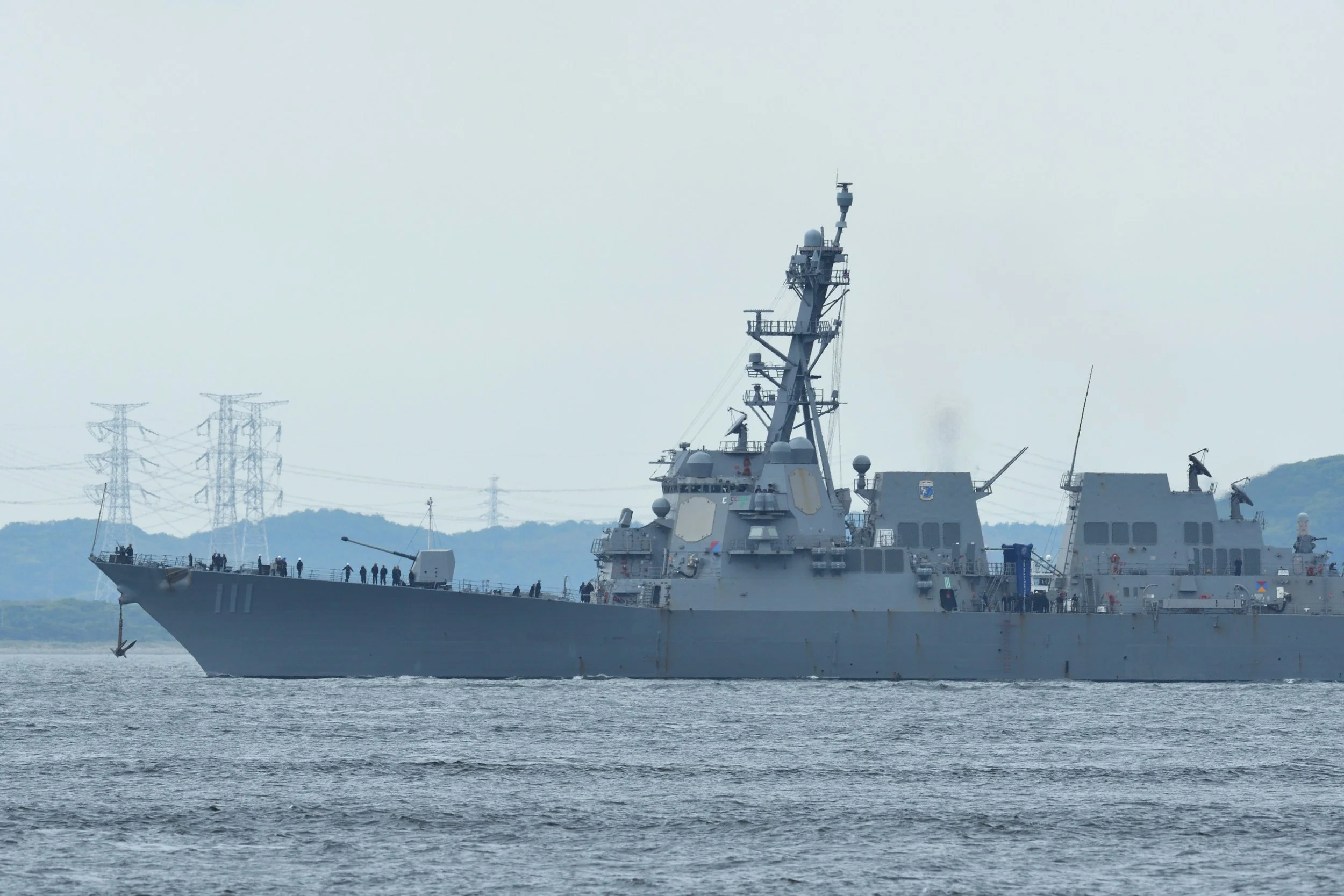 A large military Navy ship sailing on water with visible radar and communication equipment on its deck, background of hills, power lines, and a cloudy sky.