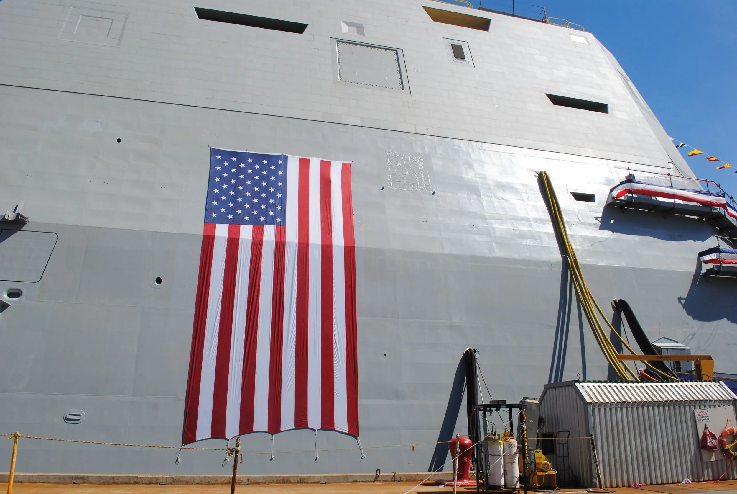 The side of a large Navy ship with an American flag hanging down. The ship's exterior is gray with several small windows and hatches. There's safety equipment and hoses at the bottom, along with a metal staircase and some machinery.