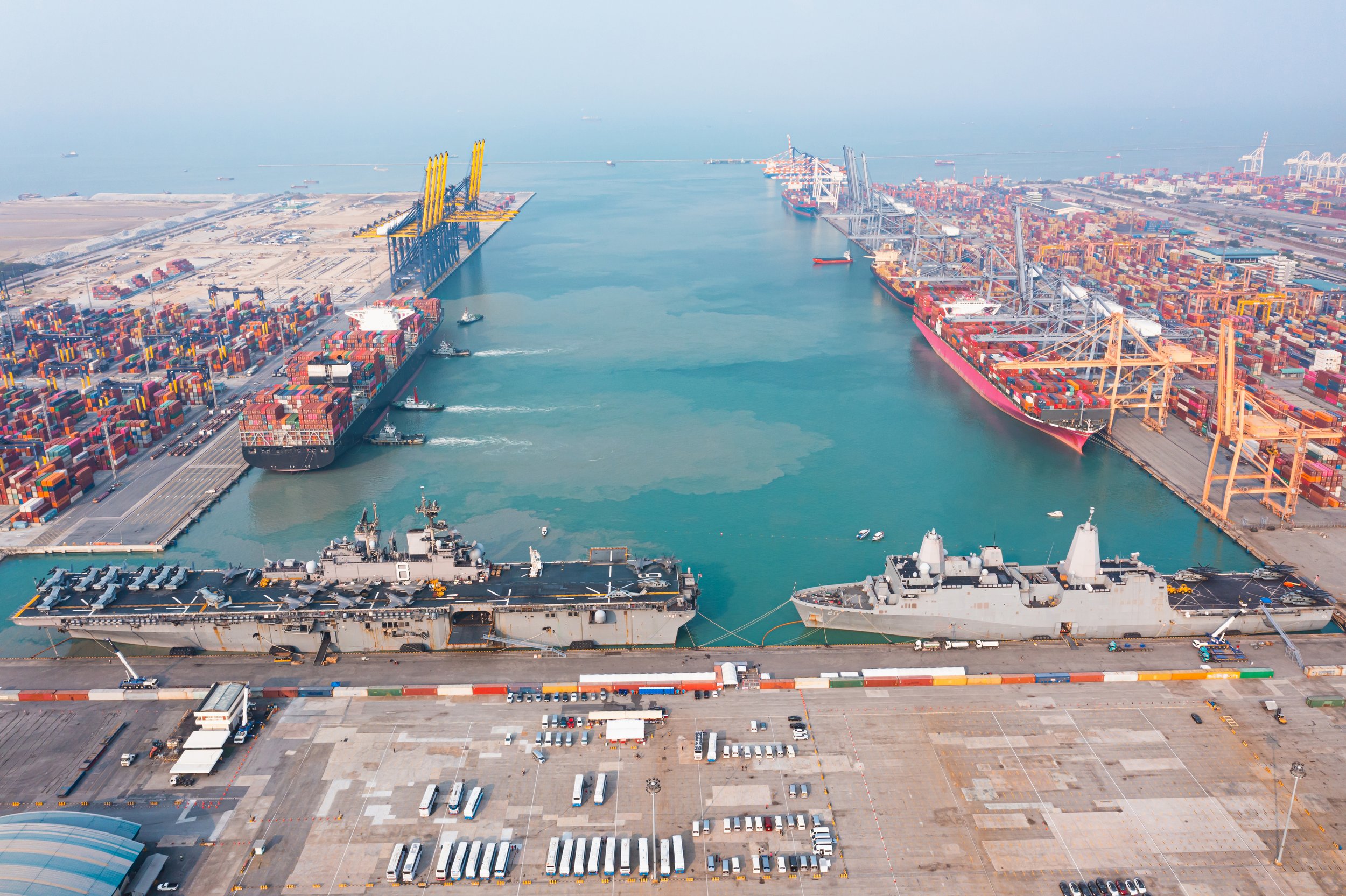Aerial view of a busy container port with Navy ships, cranes, and stacked shipping containers alongside a large cargo ship.