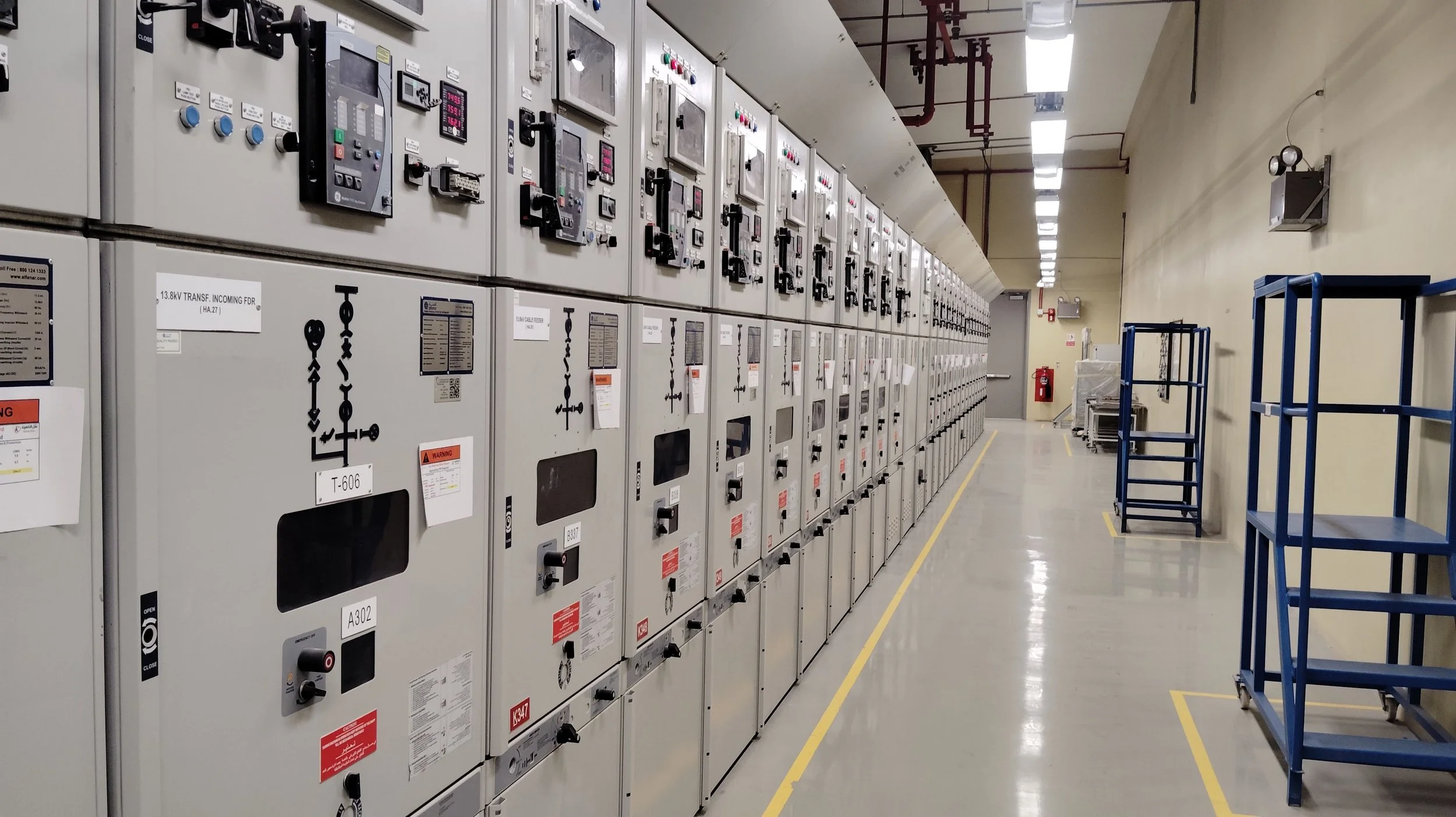 An industrial room with a long row of electrical switchgear cabinets on the left wall, a yellow line marking the floor, and a blue scaffolding trolley on the right. The room is well-lit with fluorescent lights.