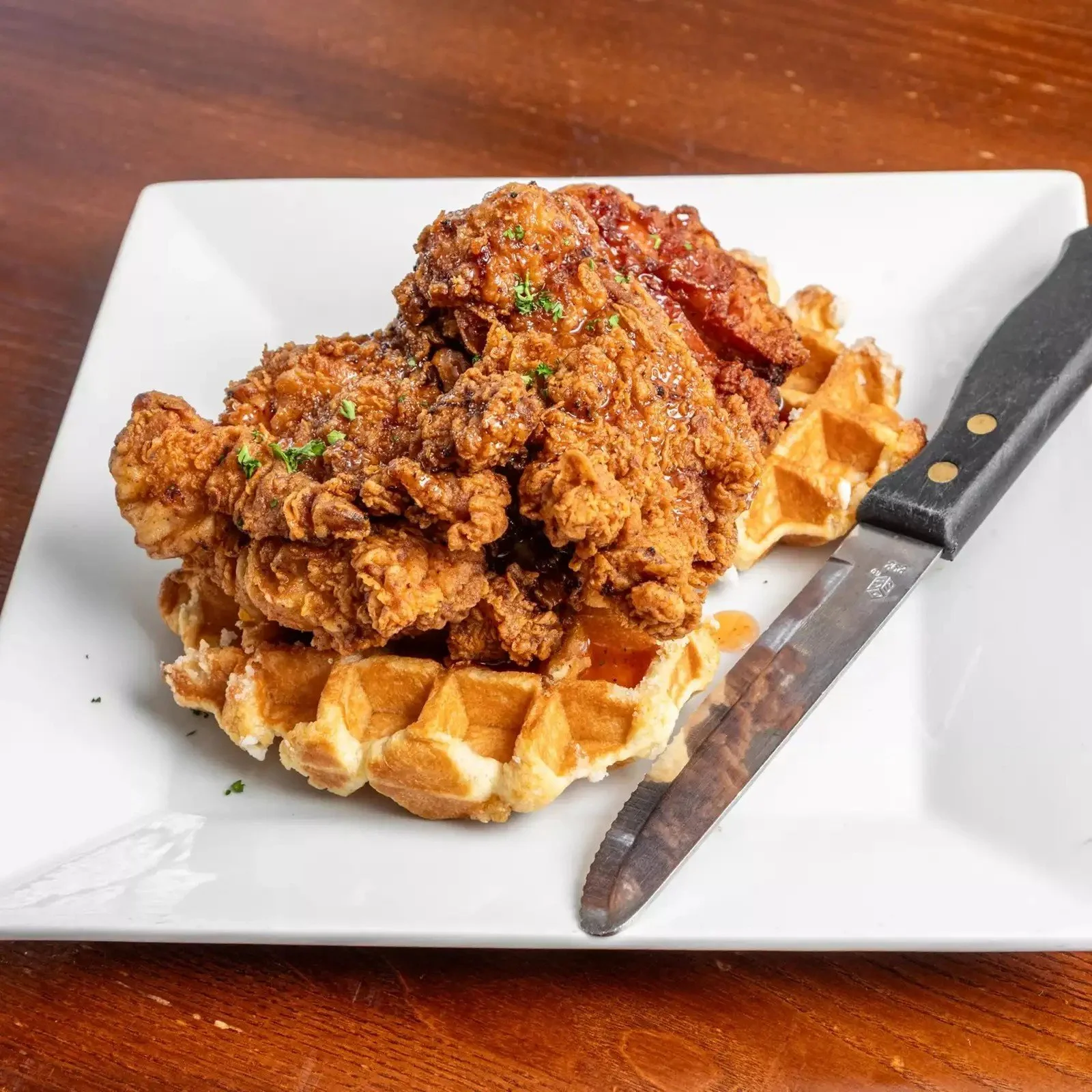 A plate with a waffle topped with fried chicken, garnished with chopped herbs, served with syrup. A knife is placed on the right side of the plate.