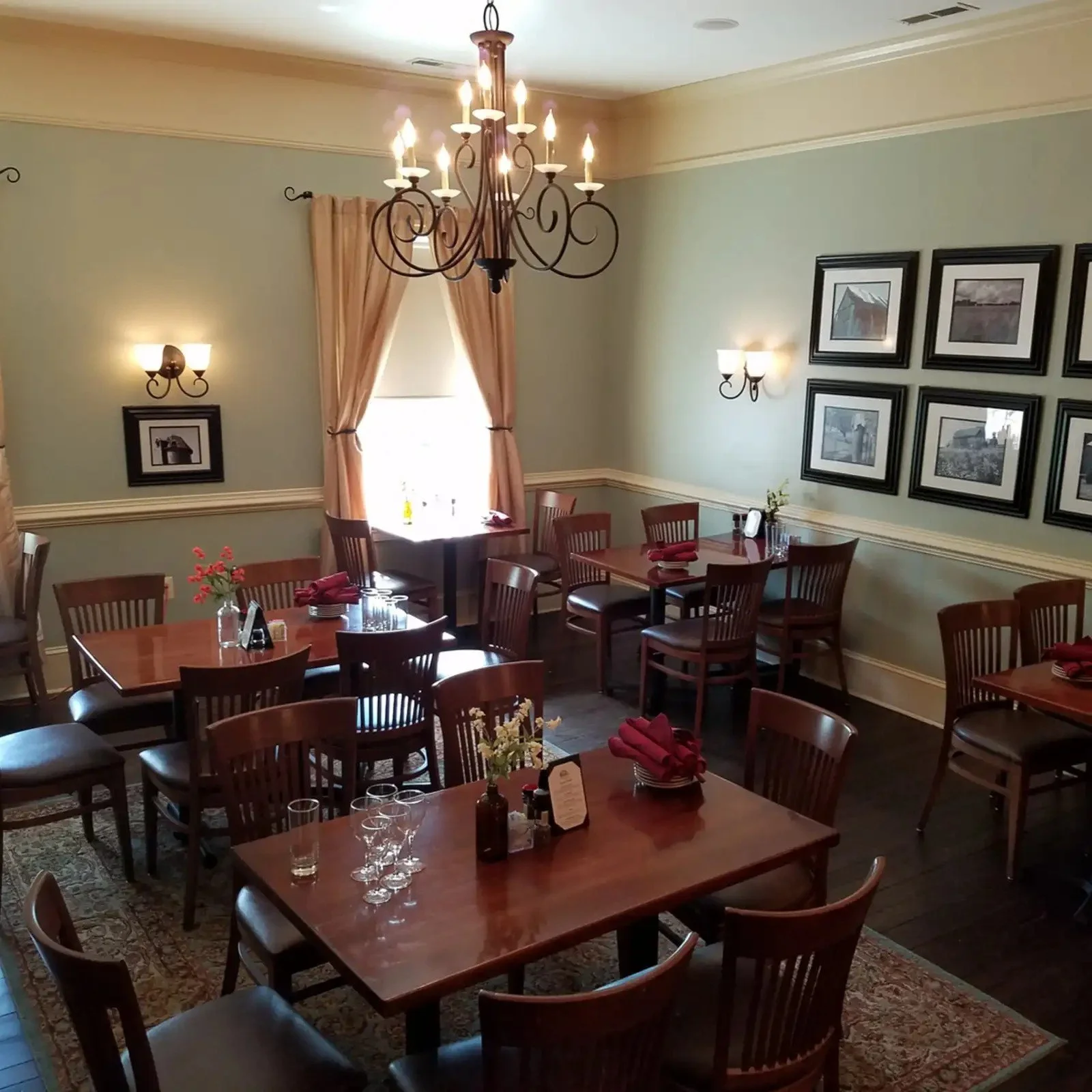 A cozy dining room with multiple wooden tables and chairs, decorated for a meal. The room features soft green walls, framed artwork, elegant wall lamps, a chandelier, and a window with beige curtains, allowing natural light to fill the space.