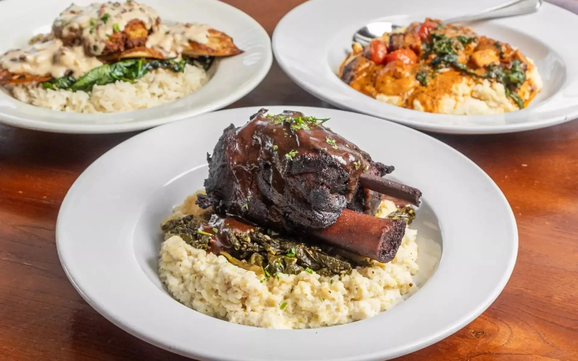 Plate with a large cooked bone-in beef short rib glazed with sauce, served over mashed potatoes with greens. Two additional plates in the background contain rice dishes with meat, vegetables, and sauces.