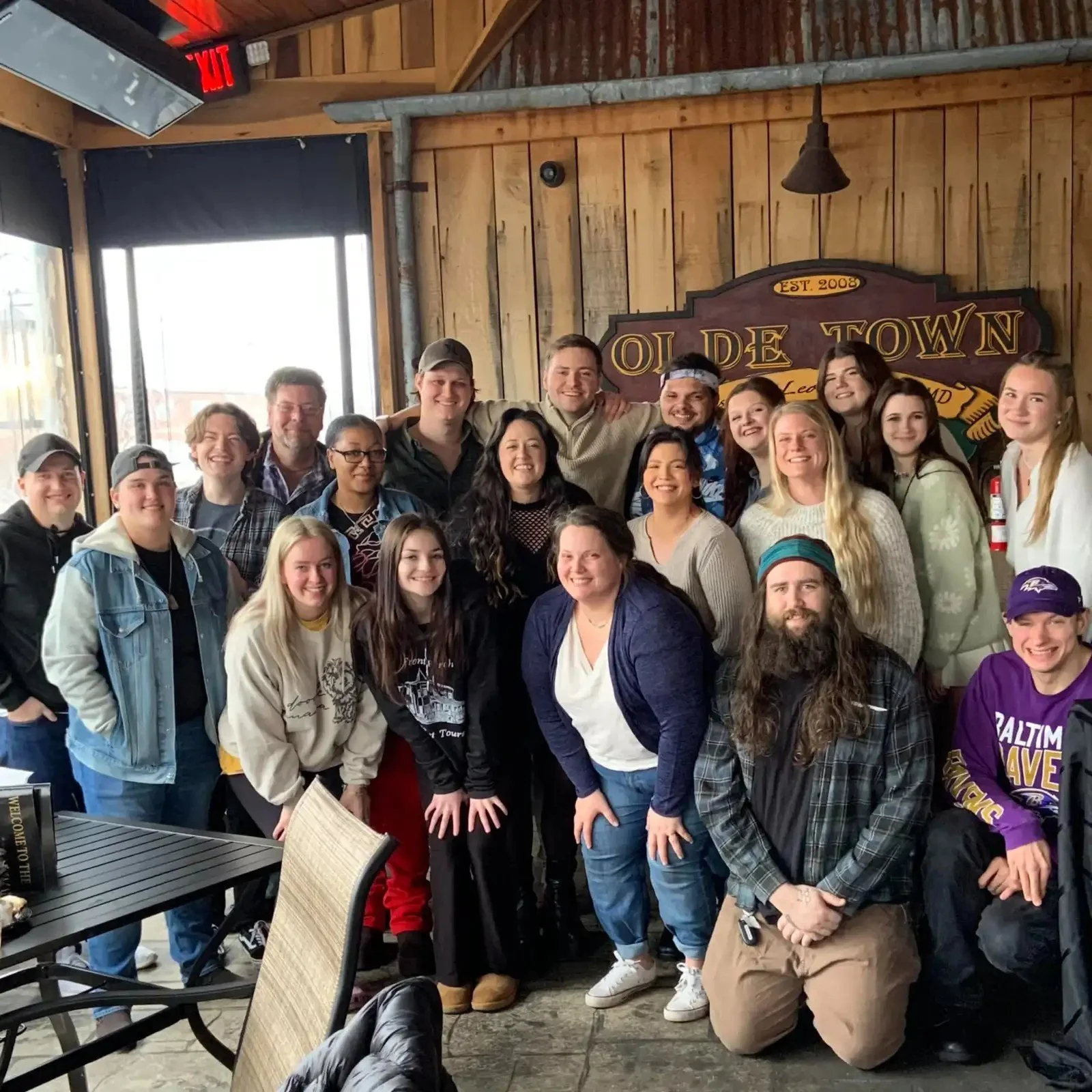 A group of about 20 diverse young adults and older individuals gathered inside a rustic wooden restaurant or café, some smiling and posing for a group photo. The background features a sign that reads 'Olde Town' with a date 'Est. 2008', and large windows letting in natural light. The setting has a cozy, cabin-like ambiance.