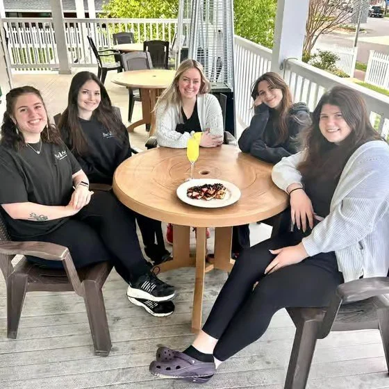 Five young women sitting around a round wooden table on an outdoor patio, smiling at the camera. The table has a plate of dessert and a drink with a yellow garnish. The patio has a white railing and other tables and chairs in the background.