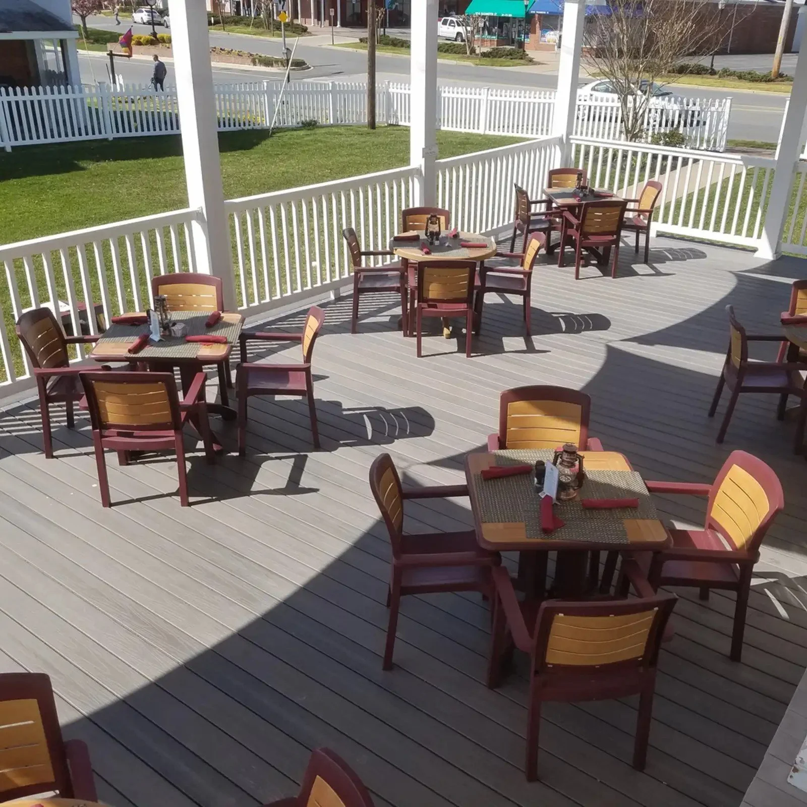 Outdoor patio with several wooden tables and chairs, set with placemats, napkins, and condiments, surrounded by a white picket fence, with a grassy yard and a street with cars and pedestrians visible in the background.