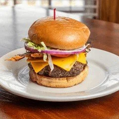 A cheeseburger with lettuce, tomato, onion, cheddar cheese, beef patty, and bacon on a bun, served on a white plate.