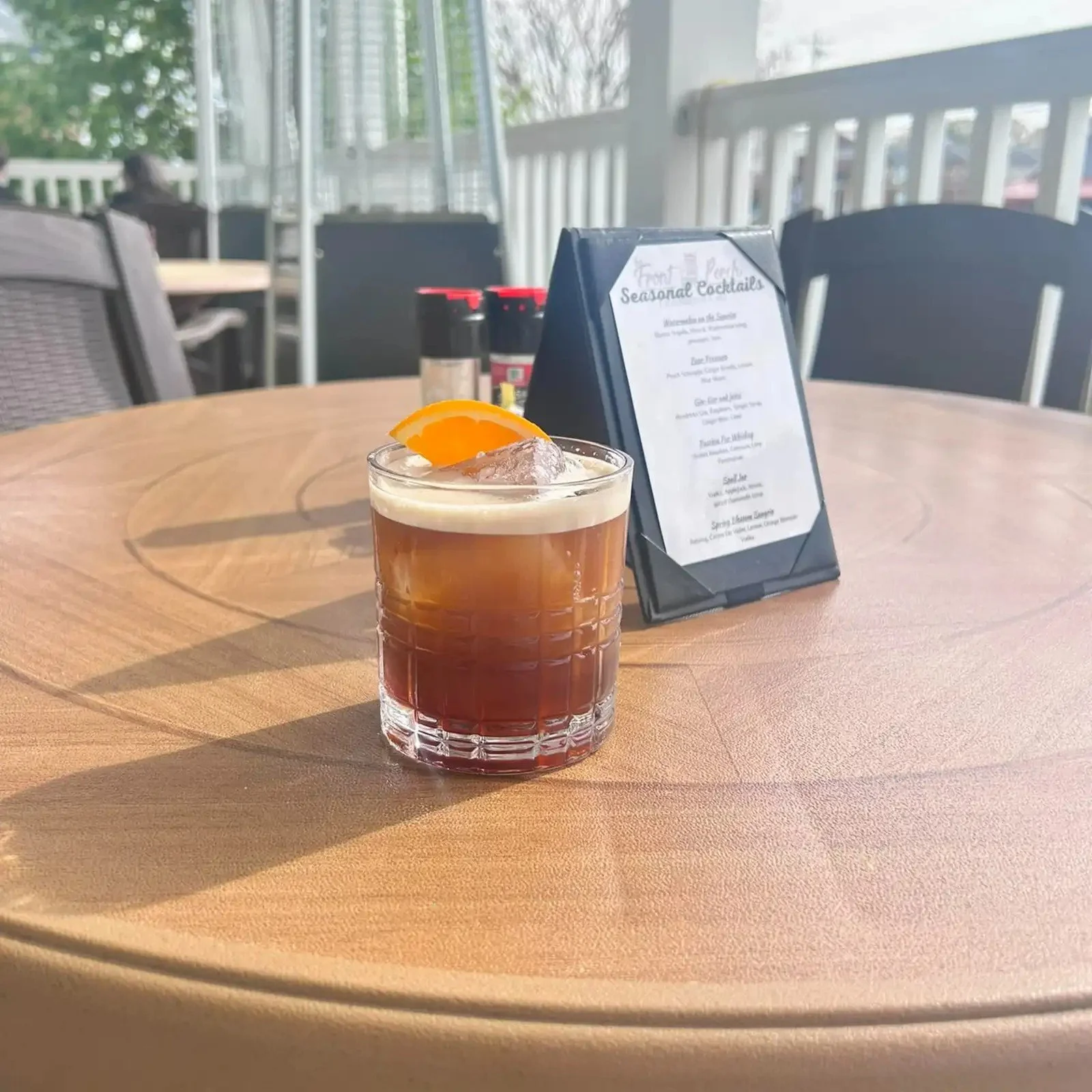 A glass of iced cocktail with an orange wedge garnish on a wooden table. In the background, a menu and condiment bottles are visible on the table, with a patio and white railing outside.