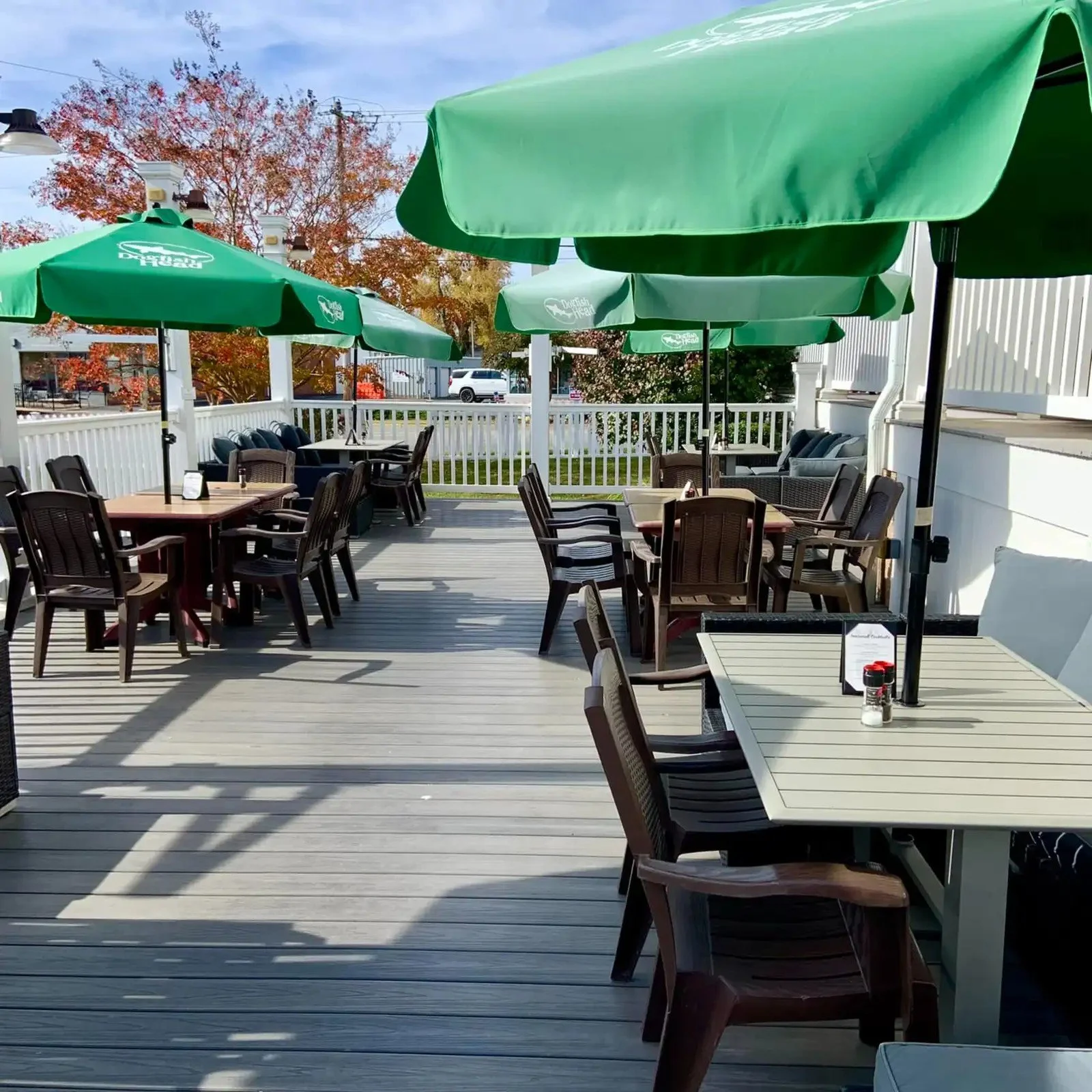 An outdoor patio with multiple tables and chairs under green umbrellas, with a white fence and trees with orange and red fall leaves in the background.