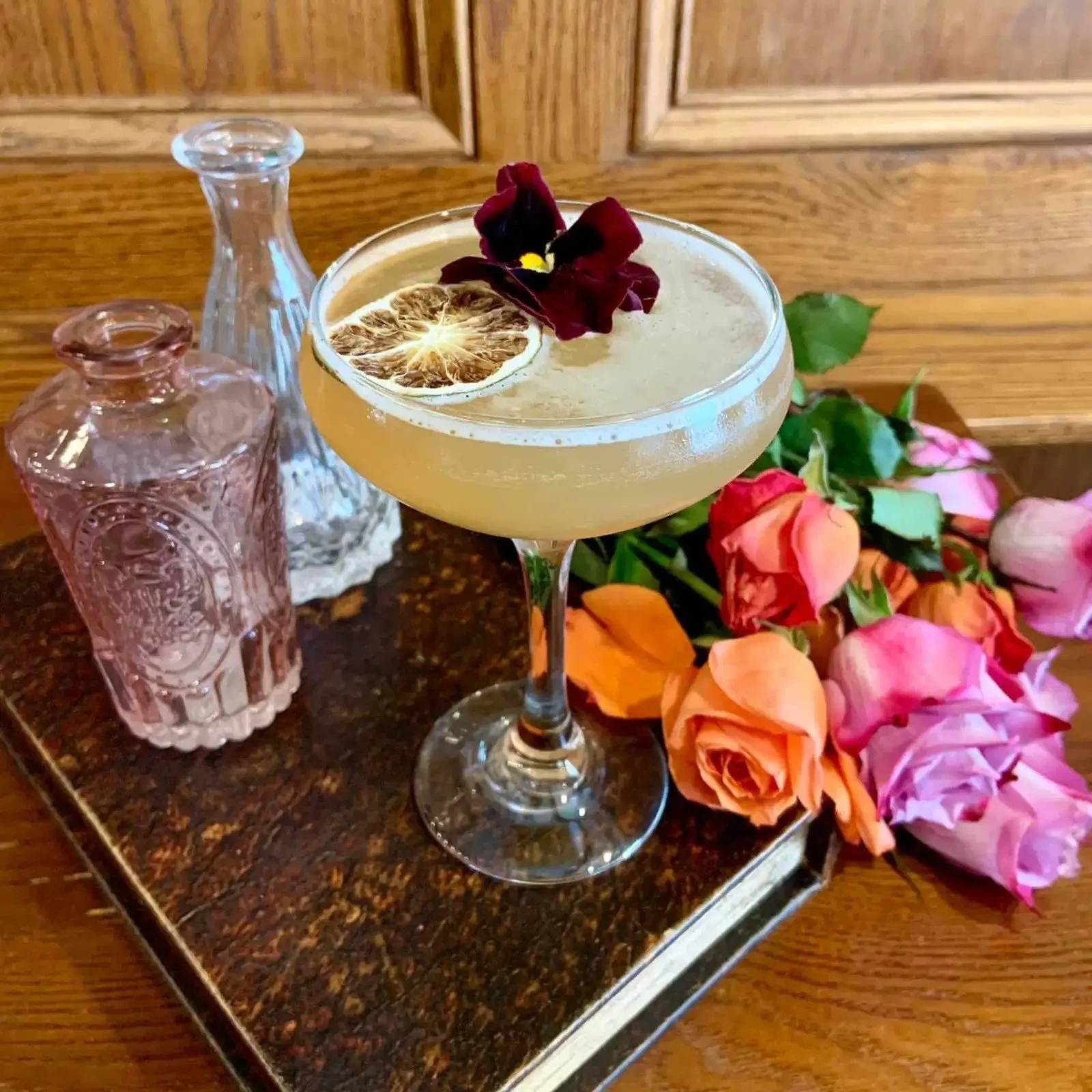 A cocktail in a stemmed glass garnished with a dried lemon slice, an edible flower, and a sprig of herbs, with pink and orange roses beside it, on a dark, textured surface.