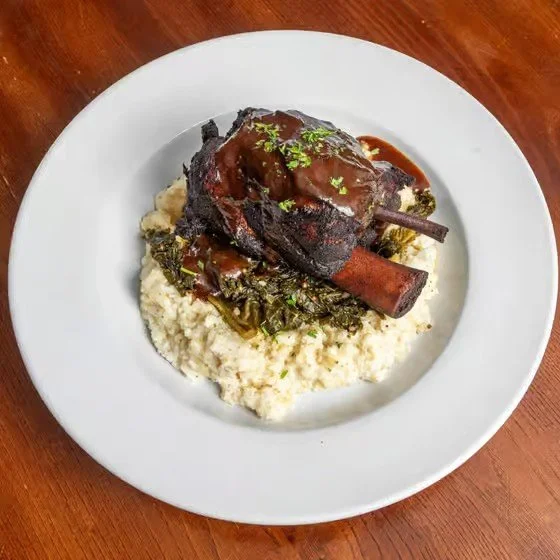 Plate with a serving of mashed potatoes topped with braised lamb shank, gravy, and green herbs.