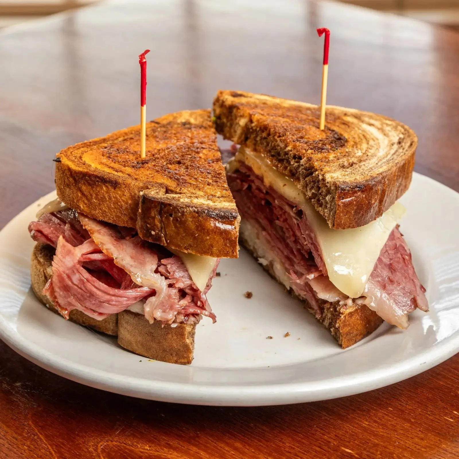 A Reuben sandwich cut in half on a white plate. The sandwich has corned beef, Swiss cheese, and sauerkraut on rye bread, with toothpicks holding it together. The plate is on a wooden table.