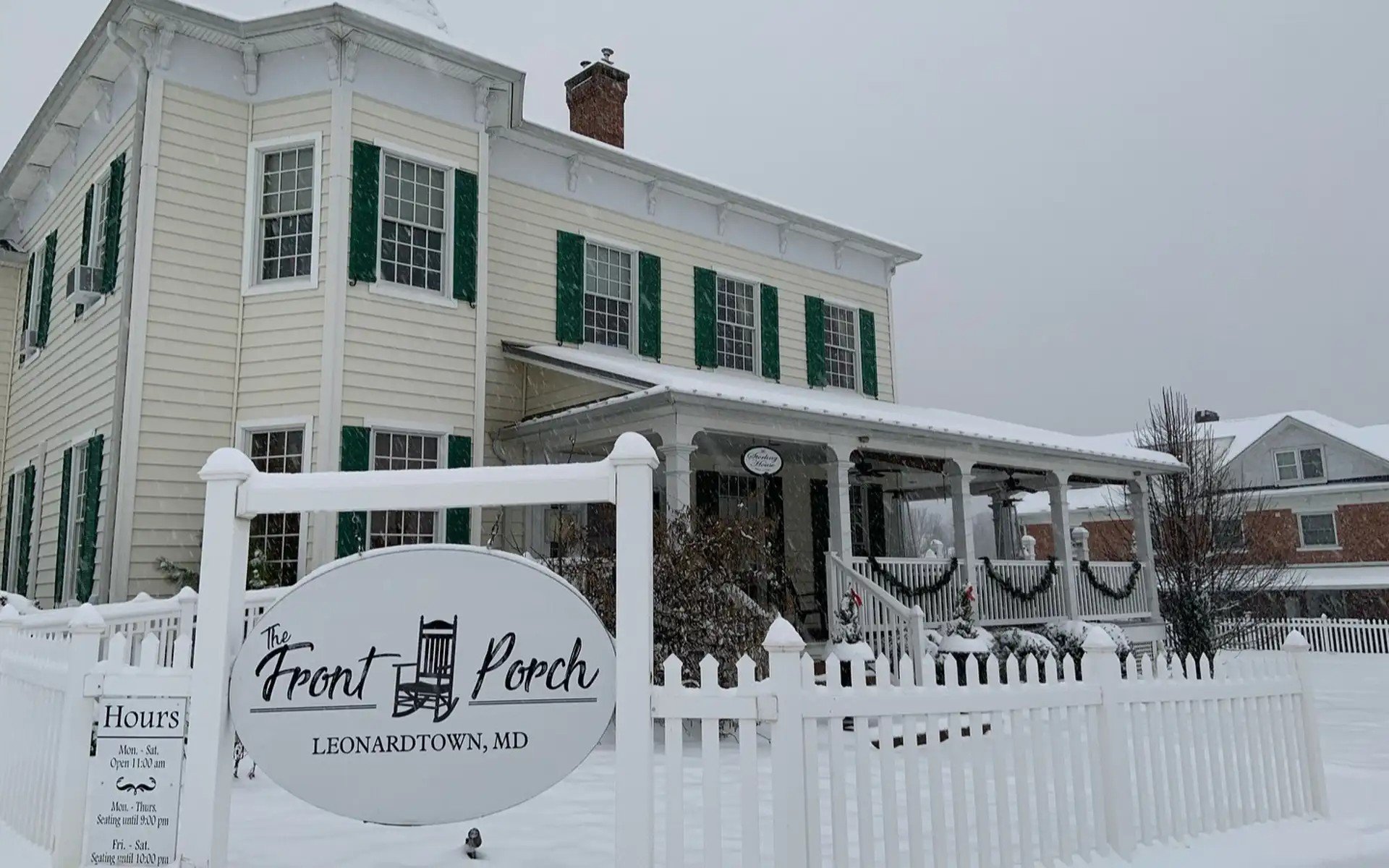 A white Victorian-style house with green shutters and snow on the roof and ground, surrounded by a white picket fence. There is a sign in front reading 'The Front Porch' with additional information about hours of operation in Leonardtown, Maryland.