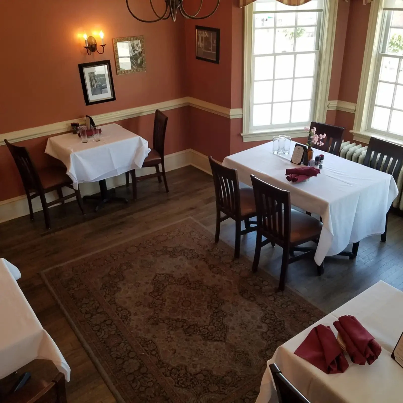 Empty dining room with three tables covered in white tablecloths and set with glasses, napkins, and condiments. The room has reddish walls, two large windows letting in natural light, framed pictures on the walls, a chandelier, and a patterned rug on wooden floor.