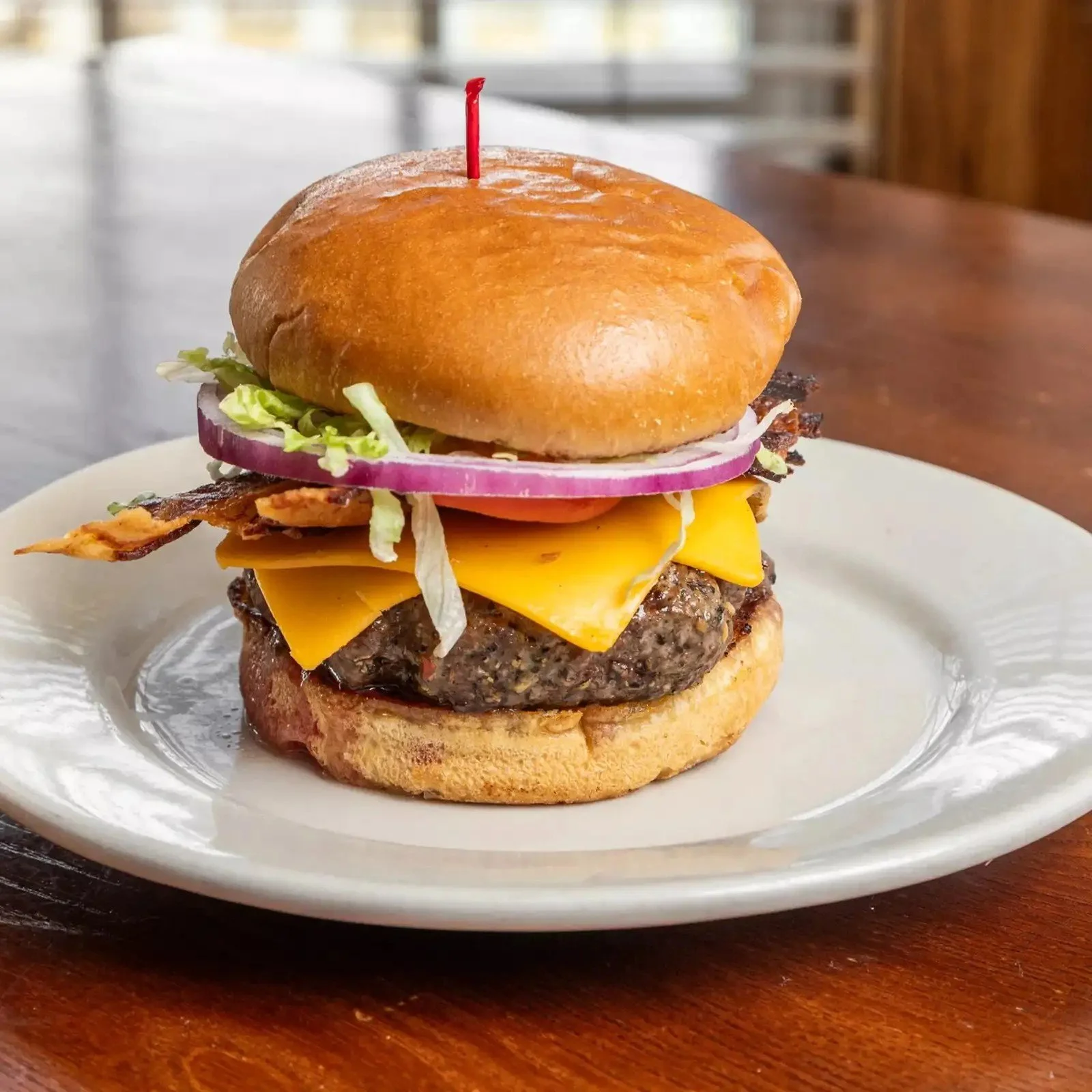 A cheeseburger with lettuce, tomato, red onion, bacon, cheddar cheese, and a beef patty on a bun, served on a white plate.