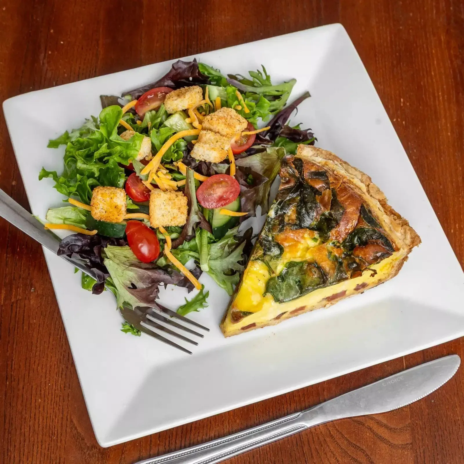 A white square plate with a salad of mixed greens, cherry tomatoes, shredded cheese, croutons, and cucumber slices, accompanied by a slice of quiche with spinach, cheese, and a golden crust, on a wooden table with a fork and knife.