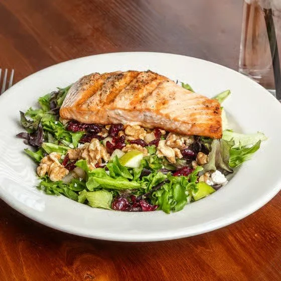 Salmon fillet on a bed of mixed greens salad with cranberries and walnuts on a white plate eating on a wooden table.