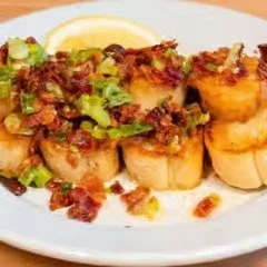 Close-up of scallops topped with bacon bits, chopped green onions, and a sauce, served on a white plate with lemon wedges and a side of vegetables.