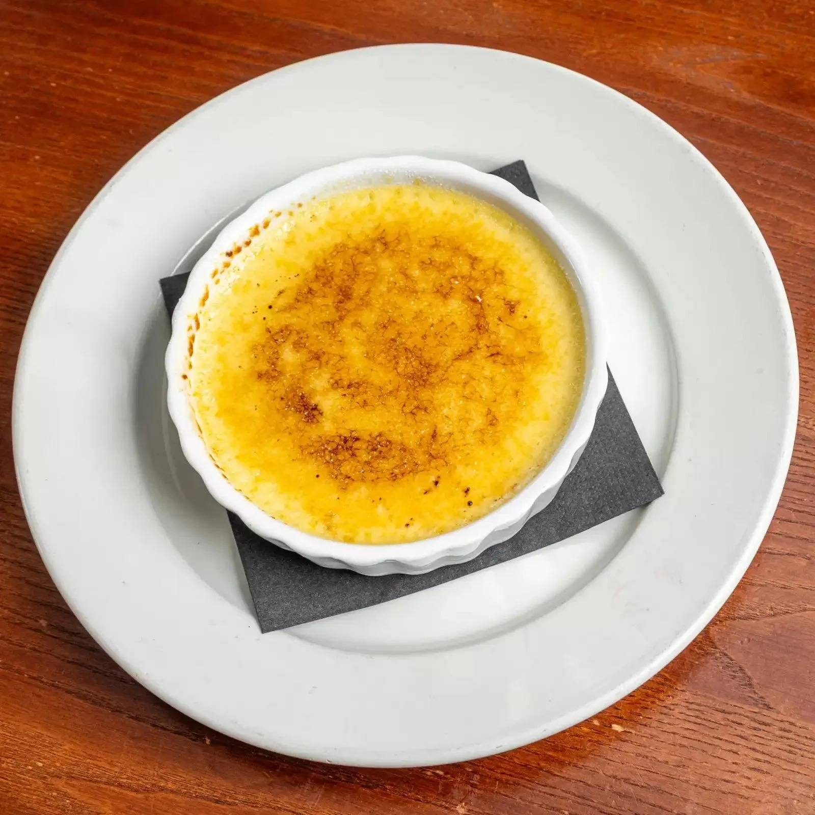 A crème brûlée dessert in a white ramekin on a white plate, placed on a wooden table.