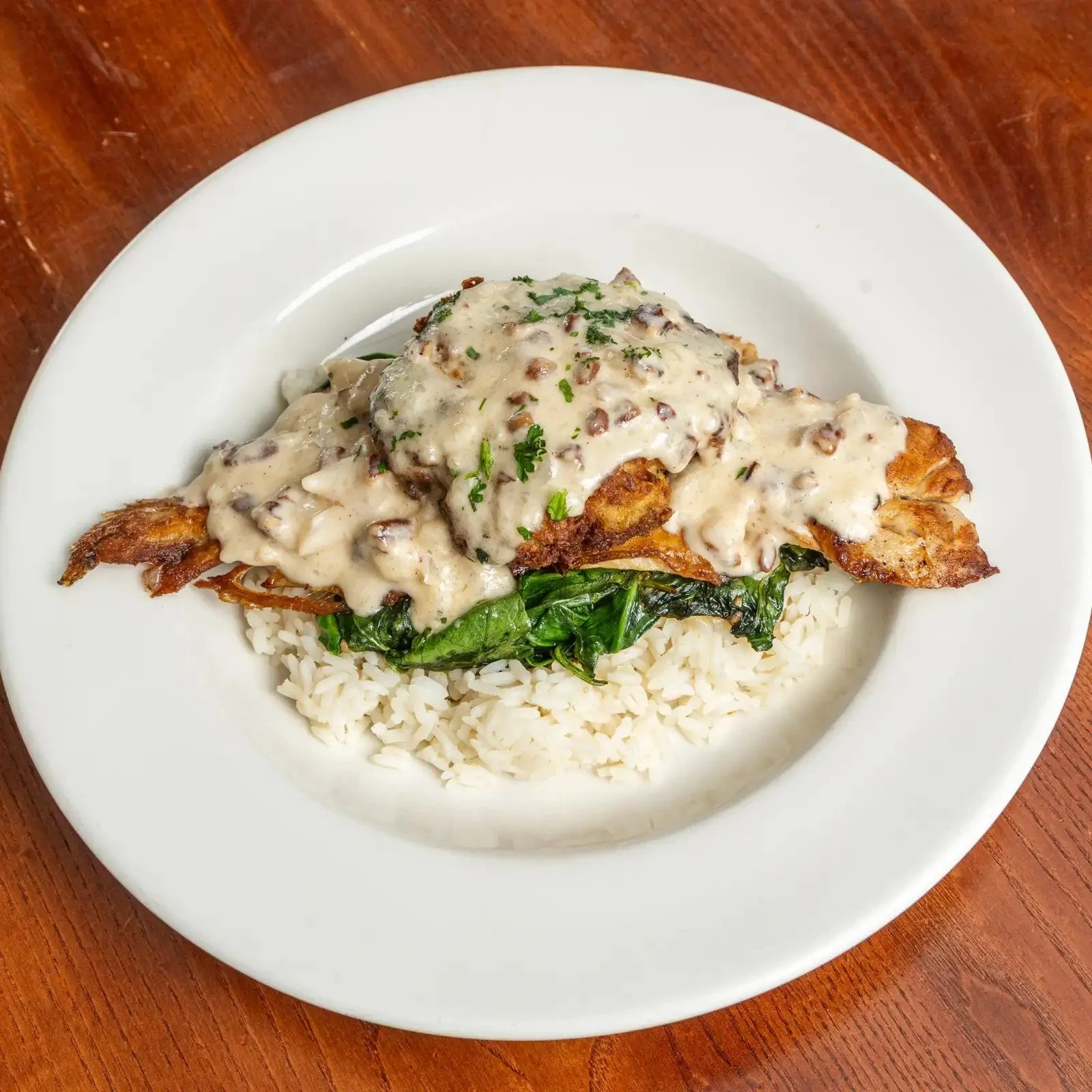 Plate with rice, cooked greens, fried fish topped with a creamy mushroom sauce garnished with parsley.