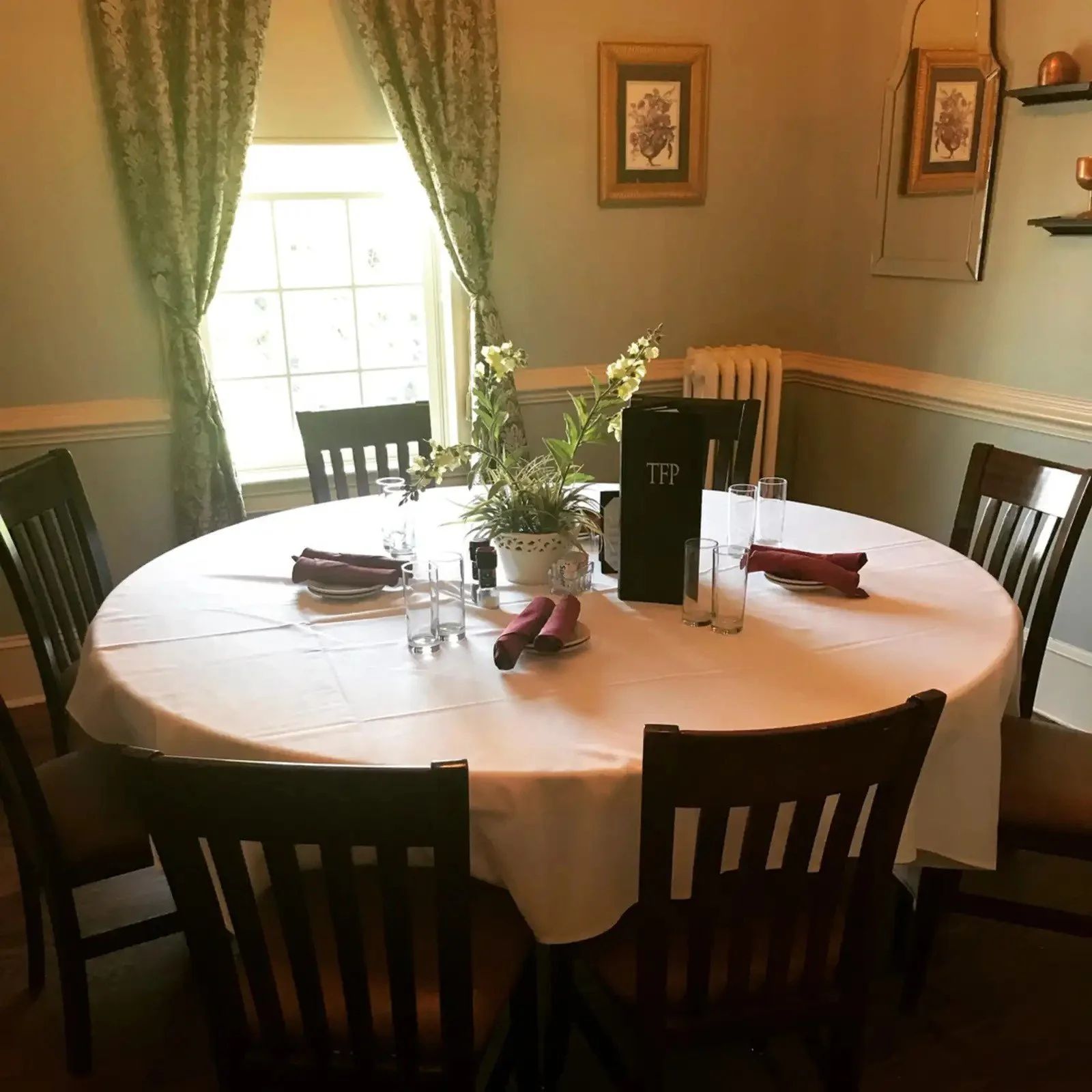 Dining room with a round table covered by a white tablecloth, set with glasses, napkins, and cutlery. A potted plant centerpiece is on the table. There are wooden chairs around the table. A window with floral curtains lets sunlight into the room. Walls are decorated with framed prints and shelves.
