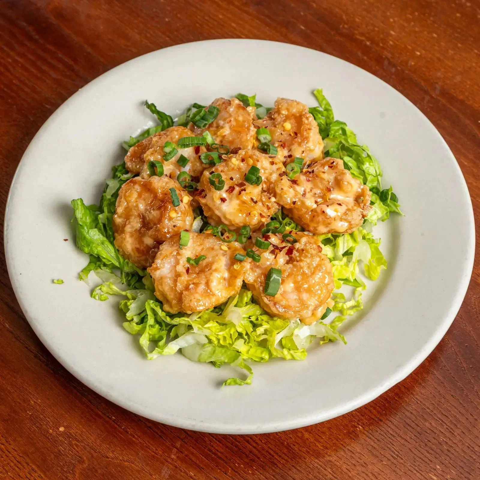 A white plate with battered fried chicken pieces topped with chopped green onions, served on shredded lettuce and sliced onions on a wooden table.