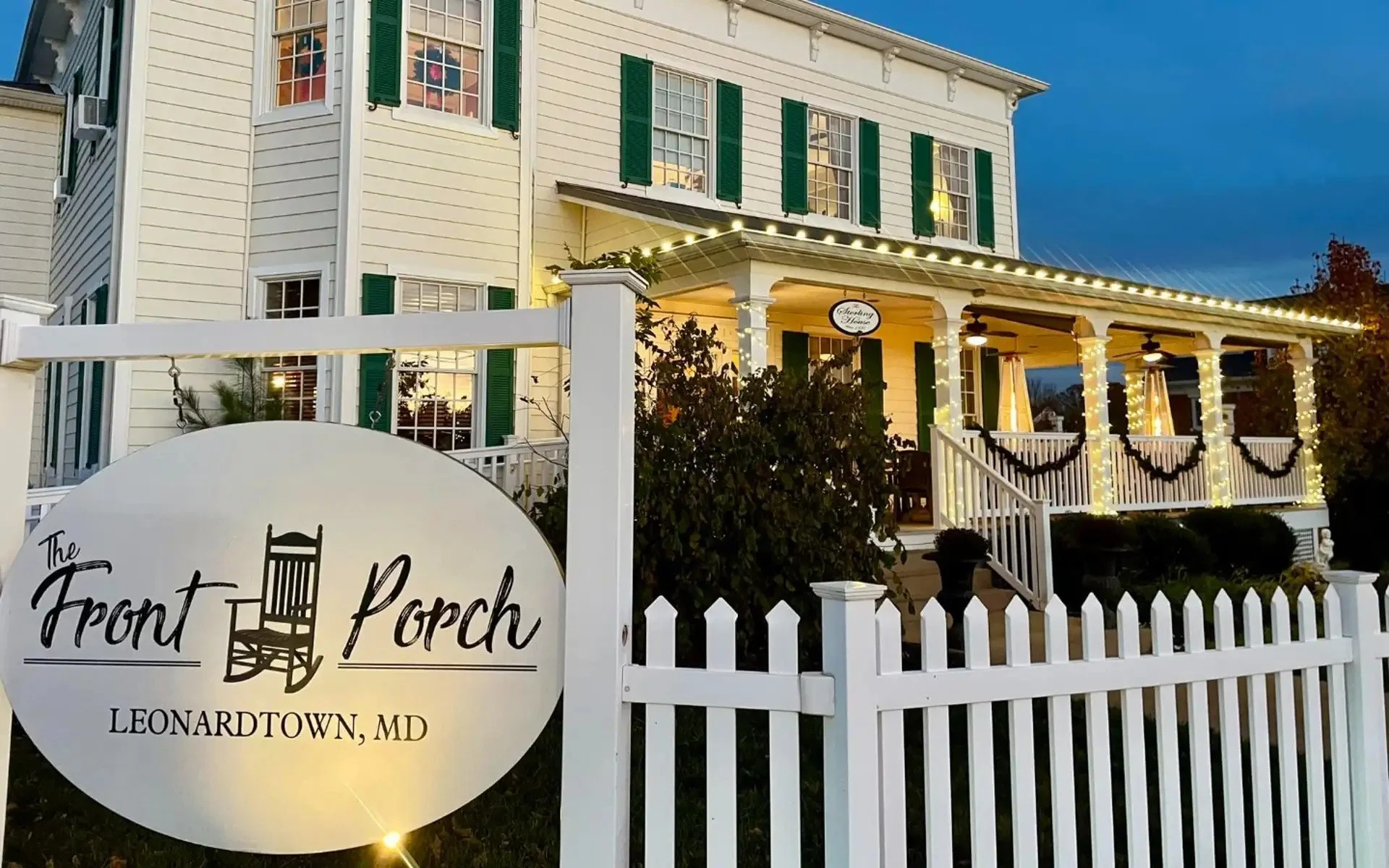 A white house with green shutters and string lights on a front porch decorated with wreaths and potted plants, in Leonardtown, Maryland, during evening hours.