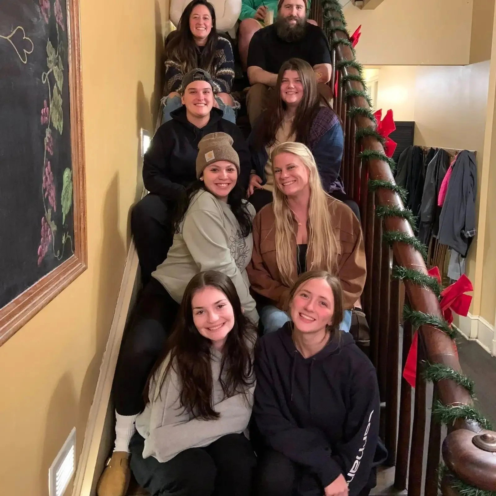 A group of people sitting on a staircase decorated with Christmas garland and red bows, smiling for a photo in an indoor setting.