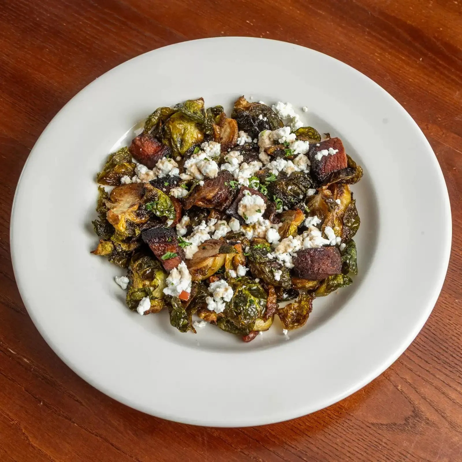 A white plate with cooked Brussels sprouts, bacon, crumbled cheese, and herbs on a wooden table.