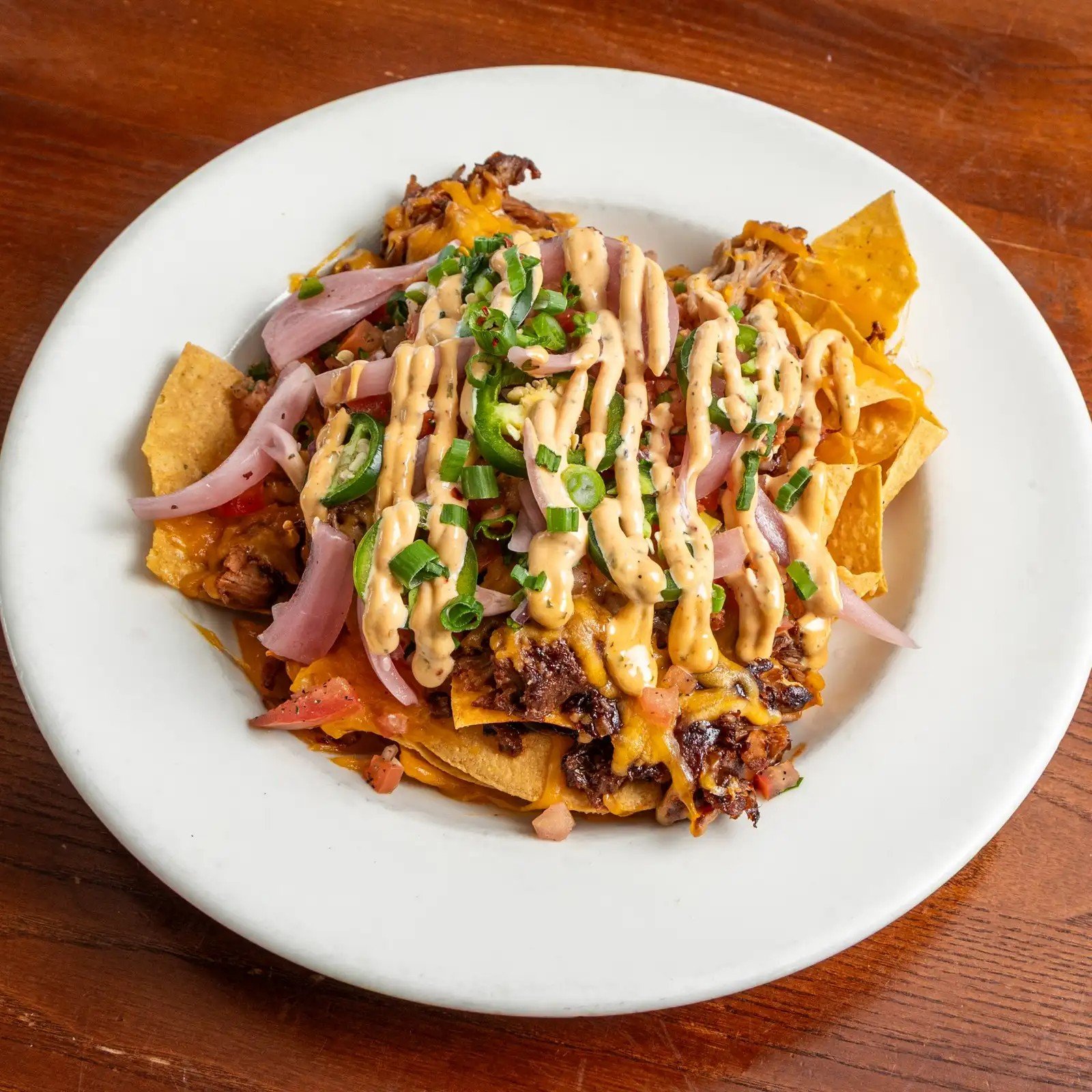 Plate of beef nachos topped with shredded cheese, chopped green onions, sliced jalapenos, sour cream, and guacamole. The nachos also include ground beef, chopped tomatoes, and chili peppers, served on a white plate on a wooden table.