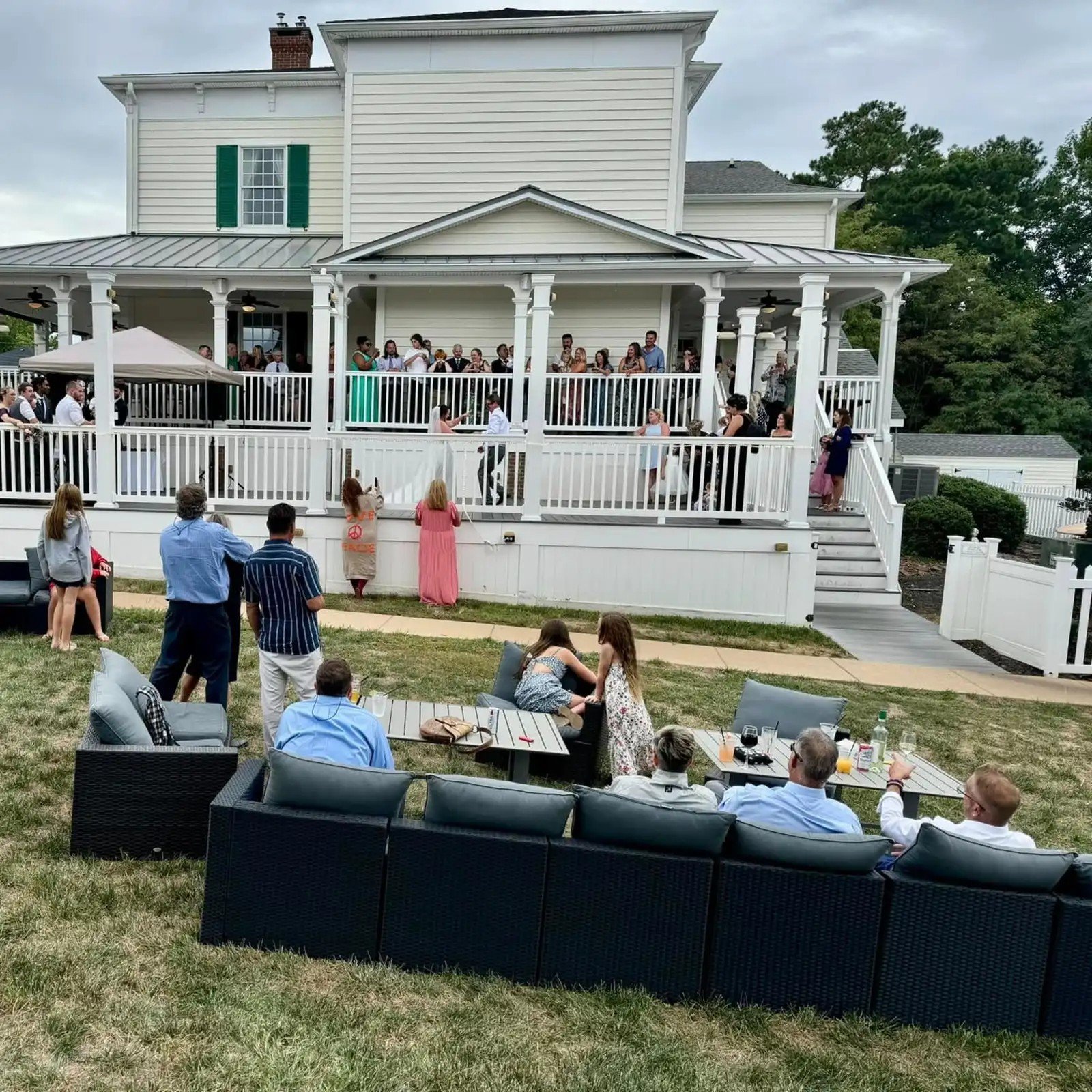 People gathering on a porch and in the yard of a large white house during a social event or celebration.