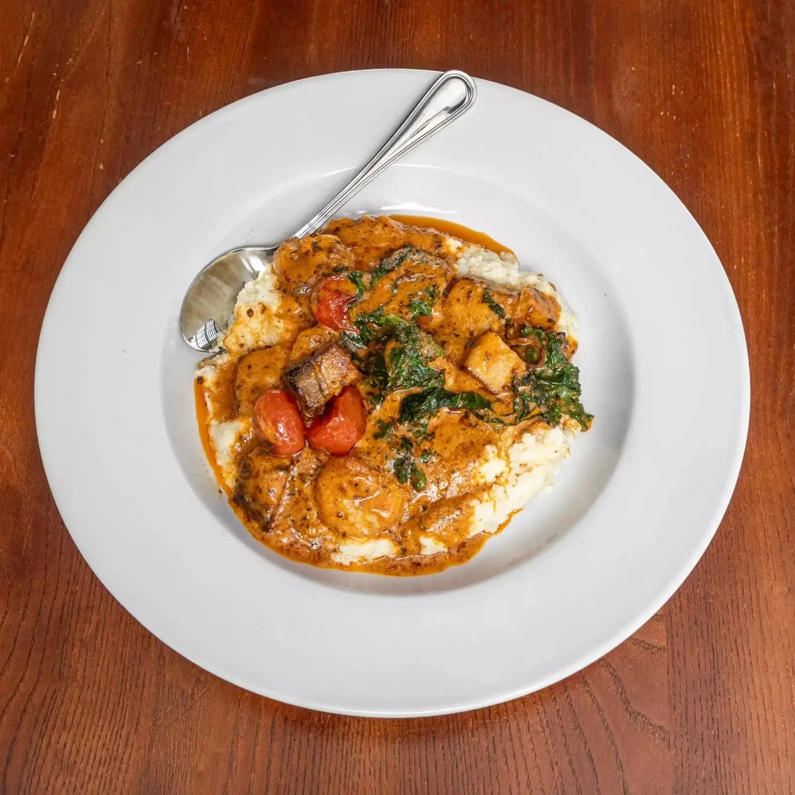 A white plate with a serving of mashed potatoes topped with beef stew, cherry tomatoes, and greens, sitting on a wooden table with a fork resting on the plate.