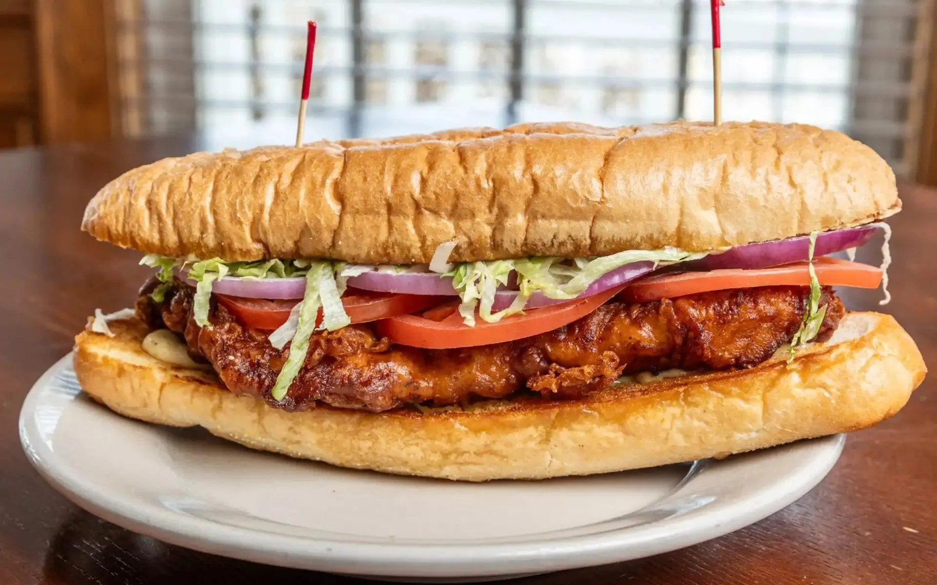 A large sandwich on a white plate with a bun, fried chicken, lettuce, tomato, and onions.