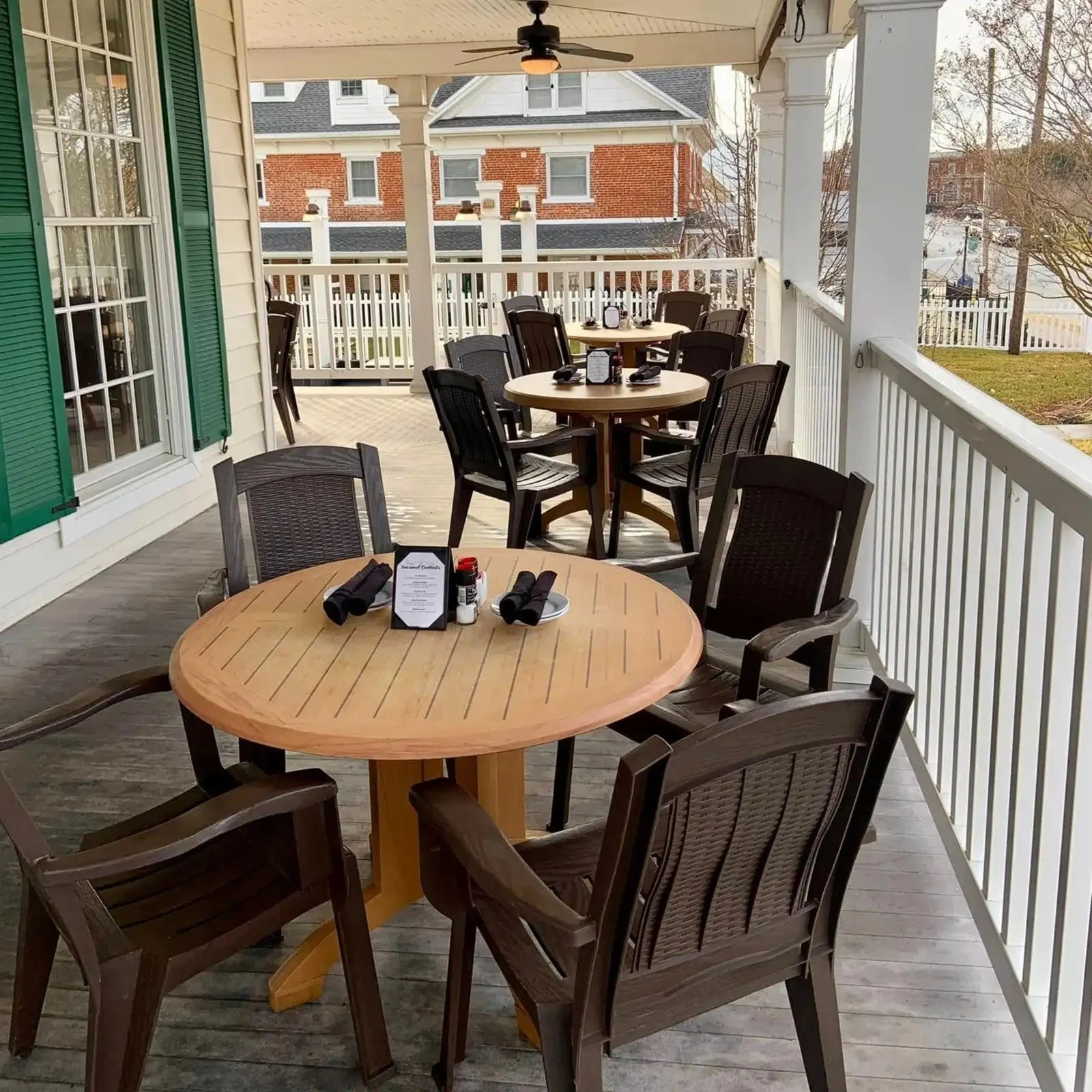 Outdoor restaurant patio with round wooden tables and dark brown plastic chairs, set with black napkins, condiments, and menus, surrounded by white railings and seen with neighboring brick buildings and leafless trees in the background.