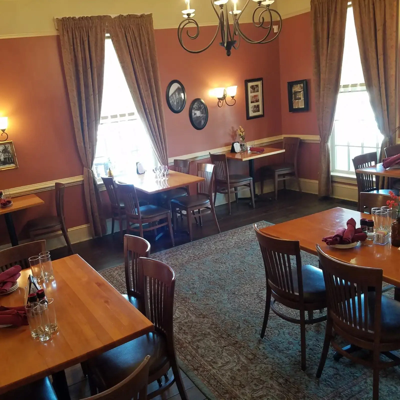 A cozy dining room with wooden tables and chairs, pink walls, framed pictures, large windows with curtains, and a chandelier.