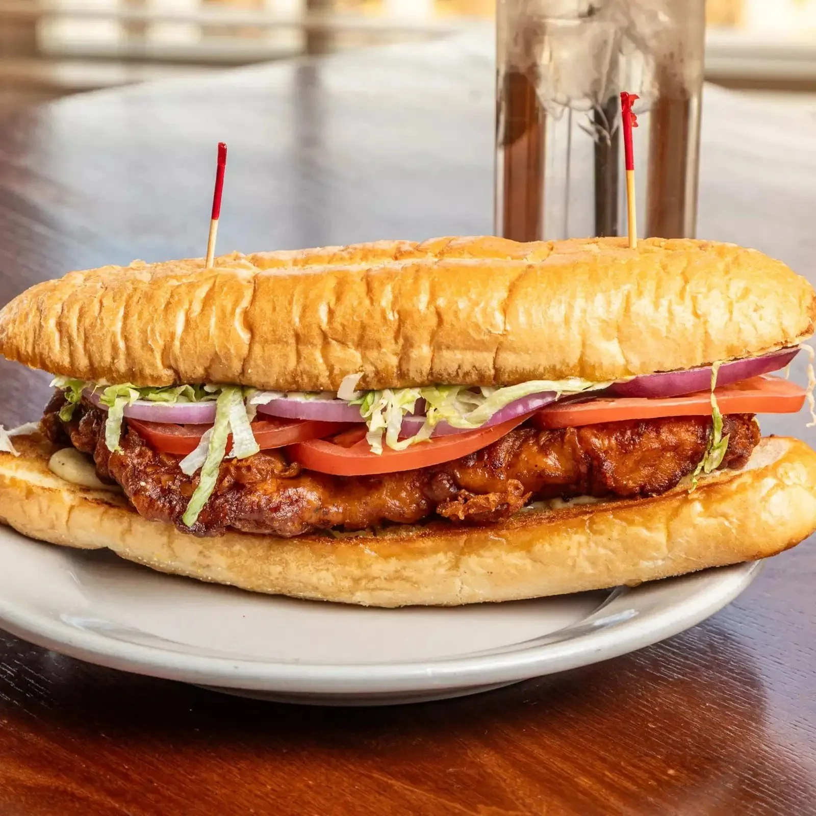 A large submarine sandwich on a white plate with fried chicken, lettuce, tomato, onion, and pickles, served on a bun with toothpicks, on a dark wooden table.