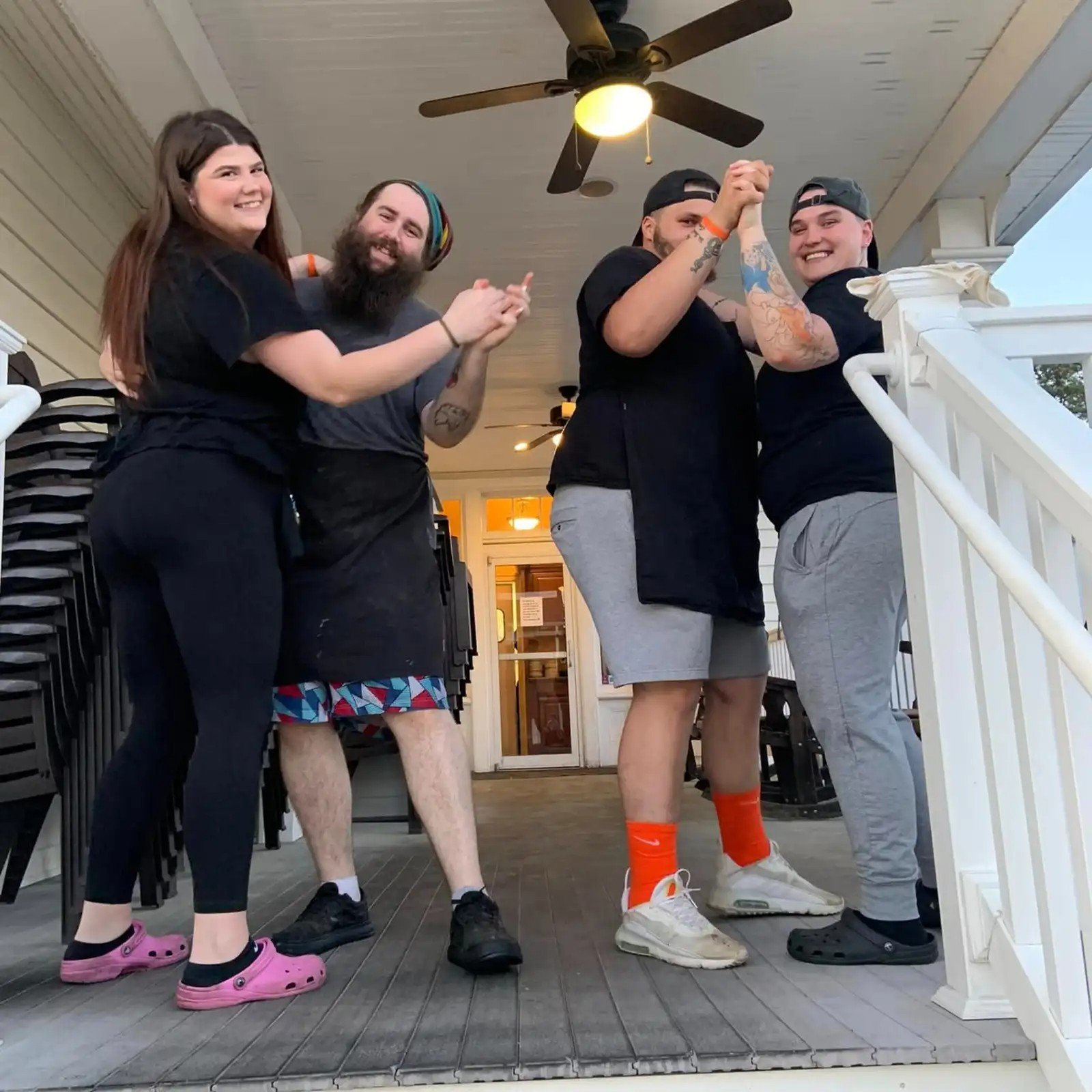 Four friends, two men and two women, are standing on a porch, smiling and showing off their muscles after arm wrestling. The woman on the left is wearing pink Crocs and black workout clothes. The man next to her has a beard, colorful headband, and is wearing black shorts. The second man has tattoos, a black cap worn backwards, and is wearing gray shorts. The woman on the right has a baseball cap, tattoos, and is wearing gray sweatpants and white sneakers. The porch has stacked chairs and a ceiling fan.