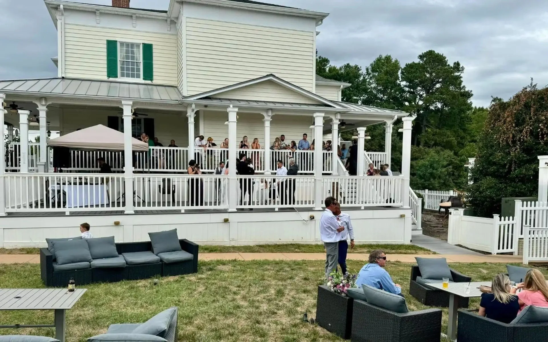People gathered on a large outdoor porch and lawn of a two-story house with cream-colored siding and white trim during a social event or gathering. Some guests are on the porch, while others are seated on outdoor furniture on the lawn. The sky is overcast, and the scene appears to be casual, with some individuals standing and chatting.